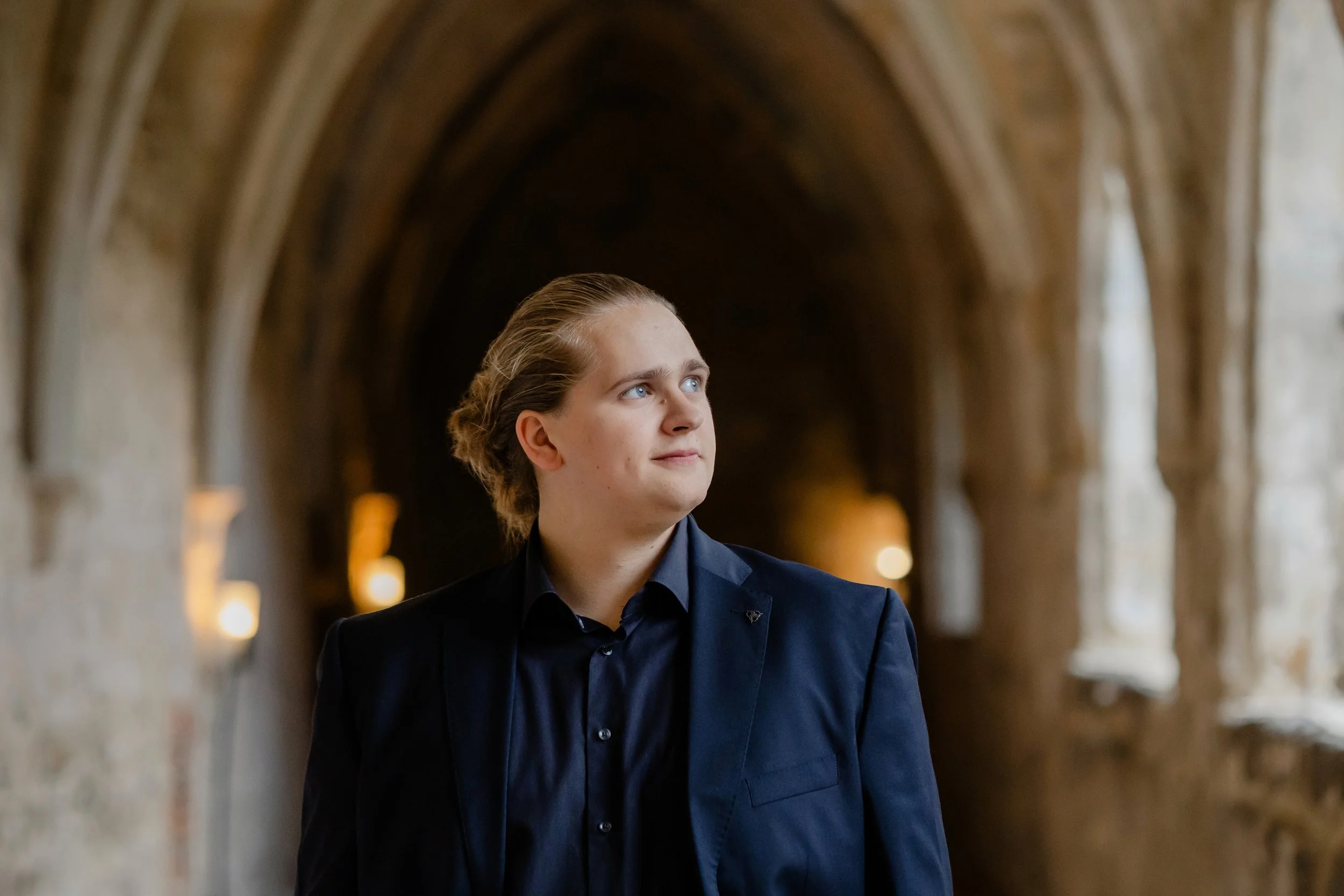 Ein junger Mann in dunklem Anzug steht in einem gotischen Flur mit gewölbter Decke, der von warmem Licht beleuchtet wird.