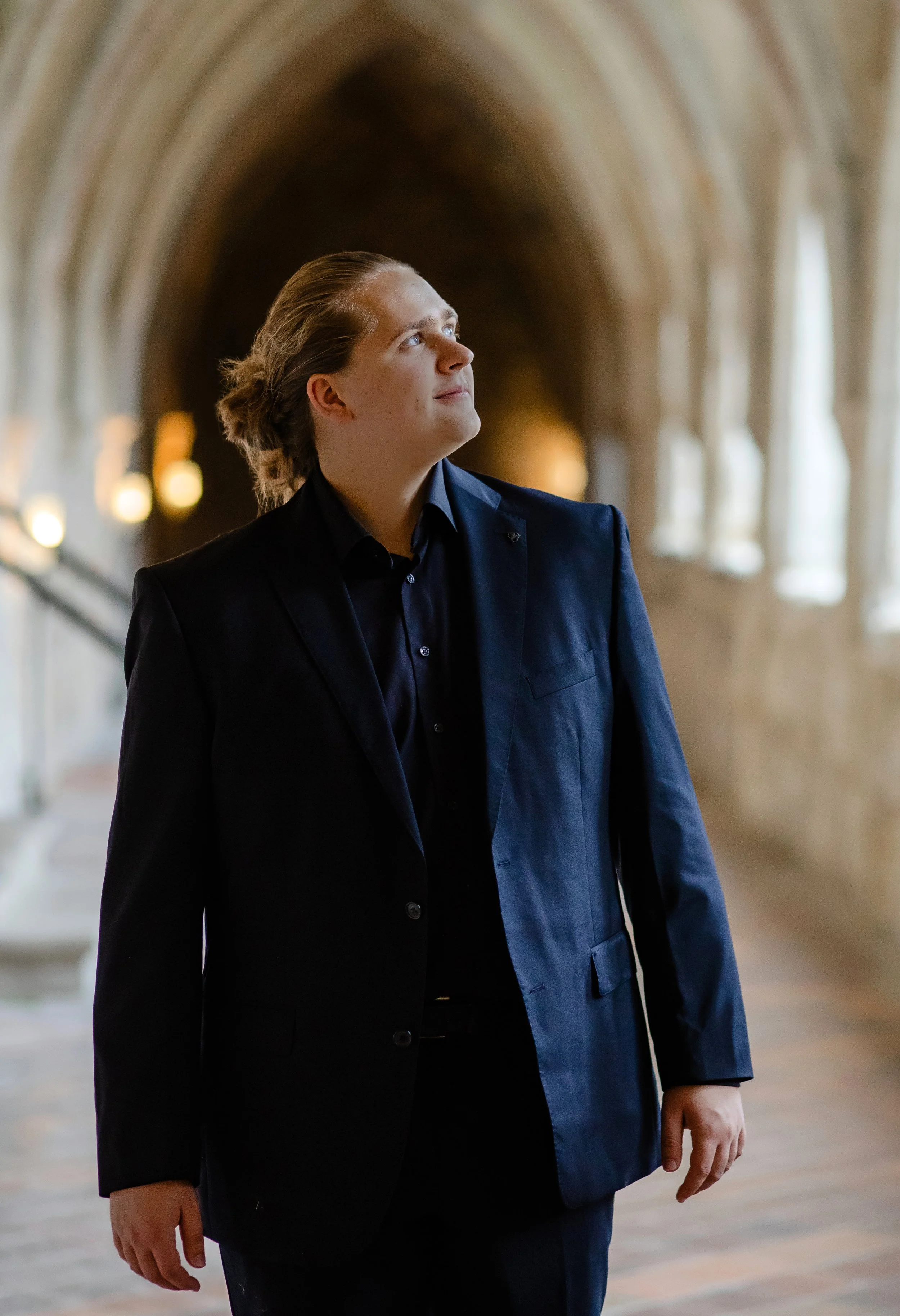 Junger Mann in dunklem Anzug steht in einem gotischen Innenraum mit hohen Bögen und Fenstern, blickt nach oben.