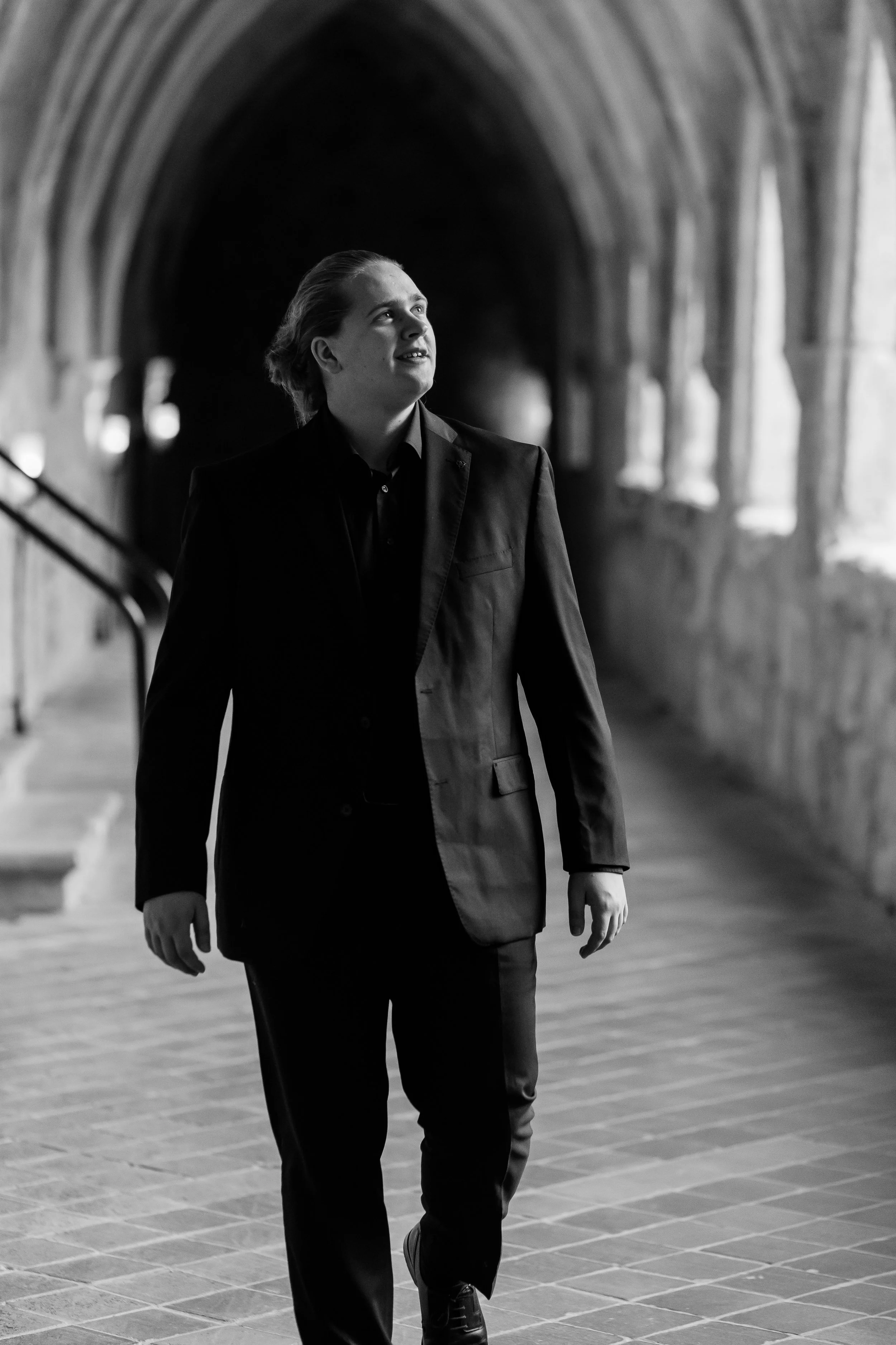 Ein junger Mann in einem schwarzen Anzug läuft durch einen langen, arched Korridor mit alten Steinen. Es ist eine Schwarz-Weiß-Aufnahme.