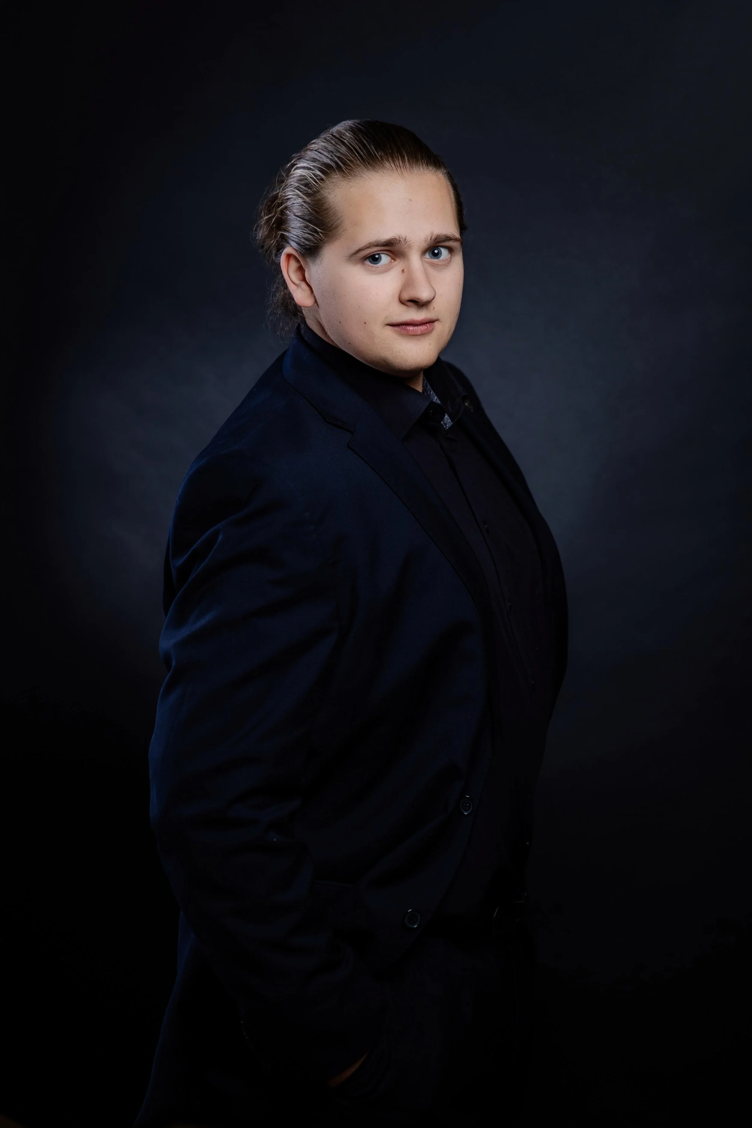 Junge Mann in dunkler Jacke vor dunklem Hintergrund, in einer professionellen Portraitaufnahme.