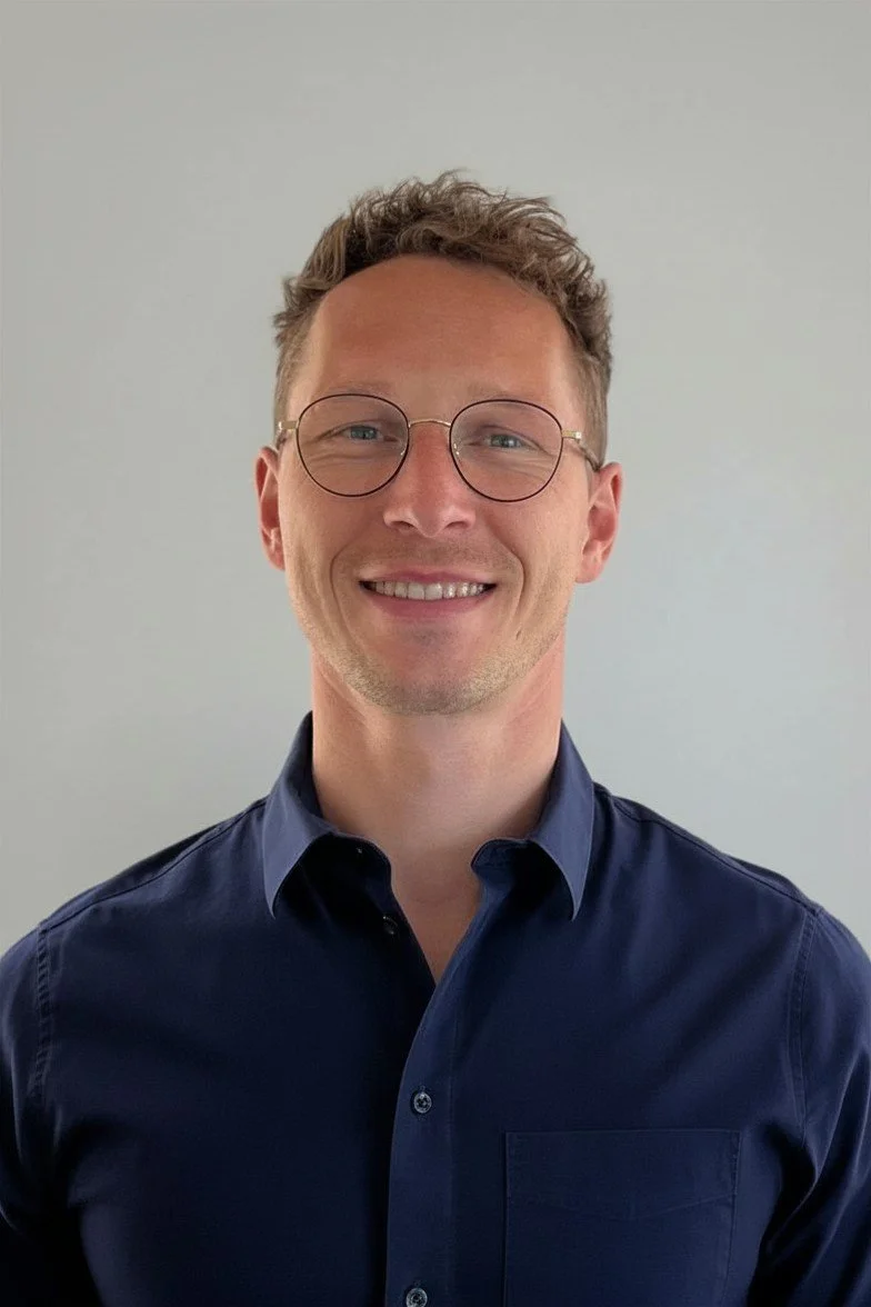 Jonas Kluge Porträt lächelt in die Kamera mit dunkelblauem Hemd und kurzen, lockigen Haaren trägt eine goldene Brille vor einem neutralen beigen Hintergrund.