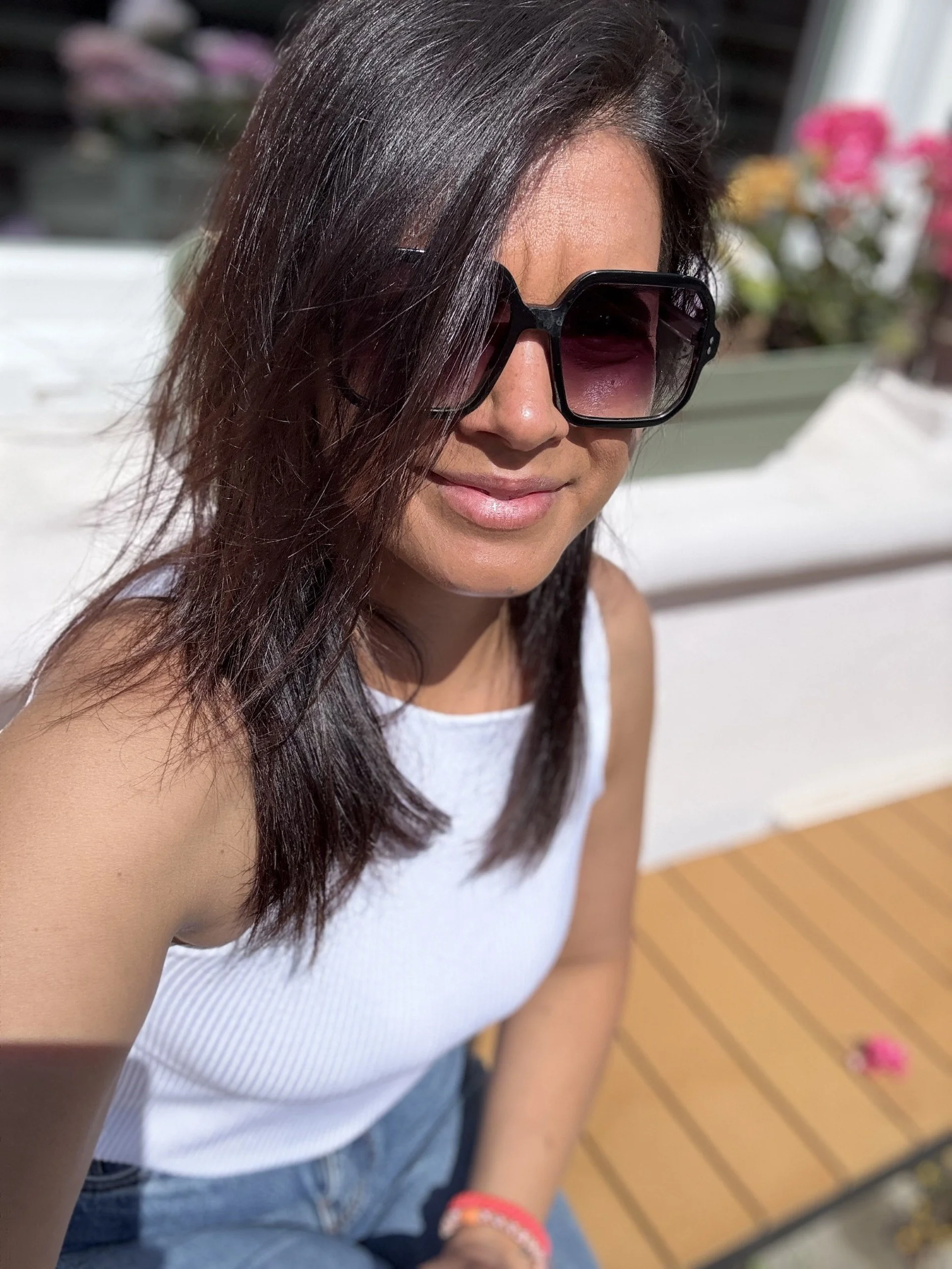 Een vrouw met donkere haar en grote zonnebril die in de zon zit, op een terras met houten vloer, terwijl ze glimlacht.