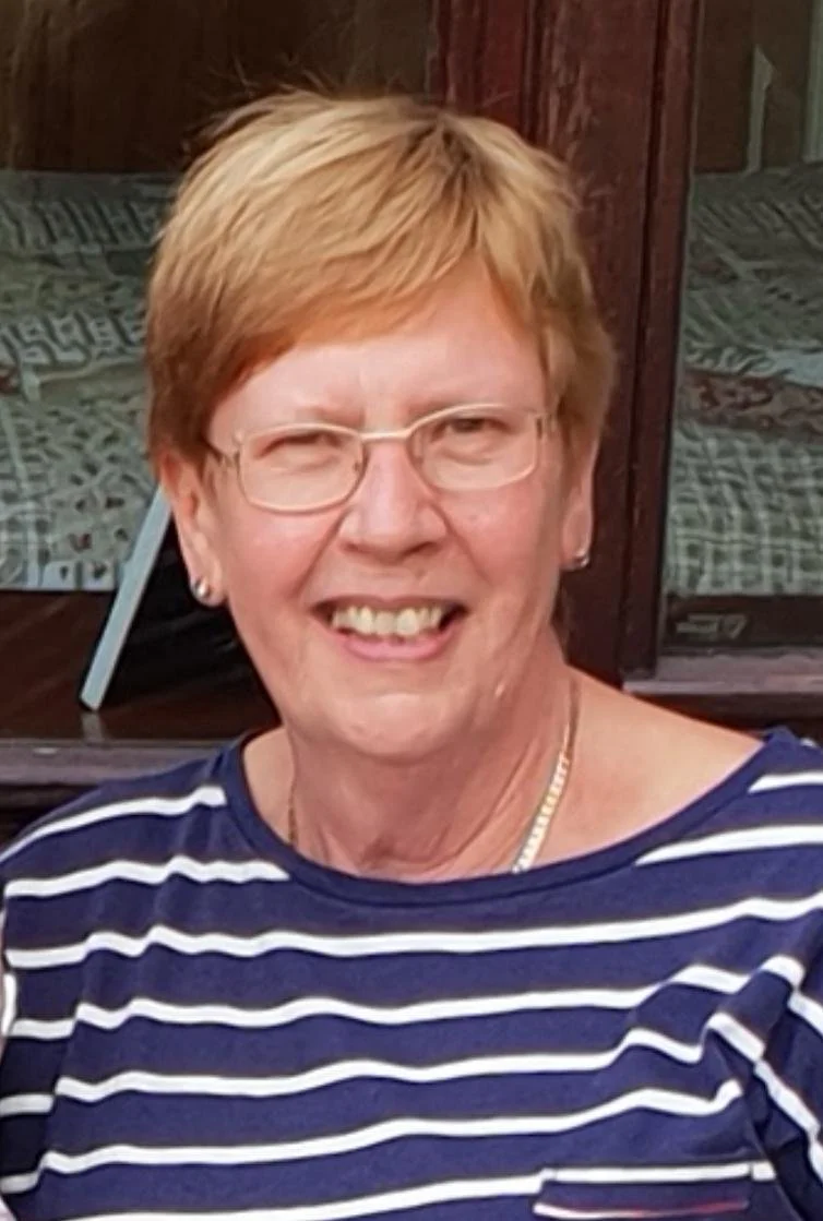 An older woman with short blonde hair, glasses, and a striped navy and white shirt smiling outside near a wooden building.