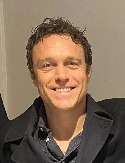 Close-up of a smiling man with curly brown hair, wearing a dark jacket over a collared shirt.