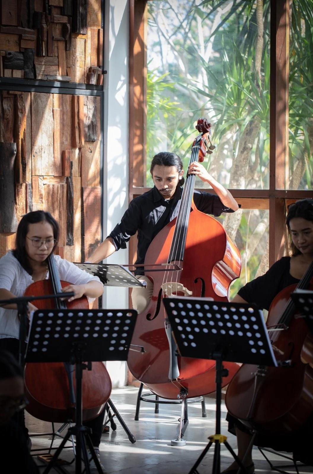 Musicians playing string instruments, including a double bass and cellos, during a rehearsal or performance in a room with large windows and wooden wall decor.