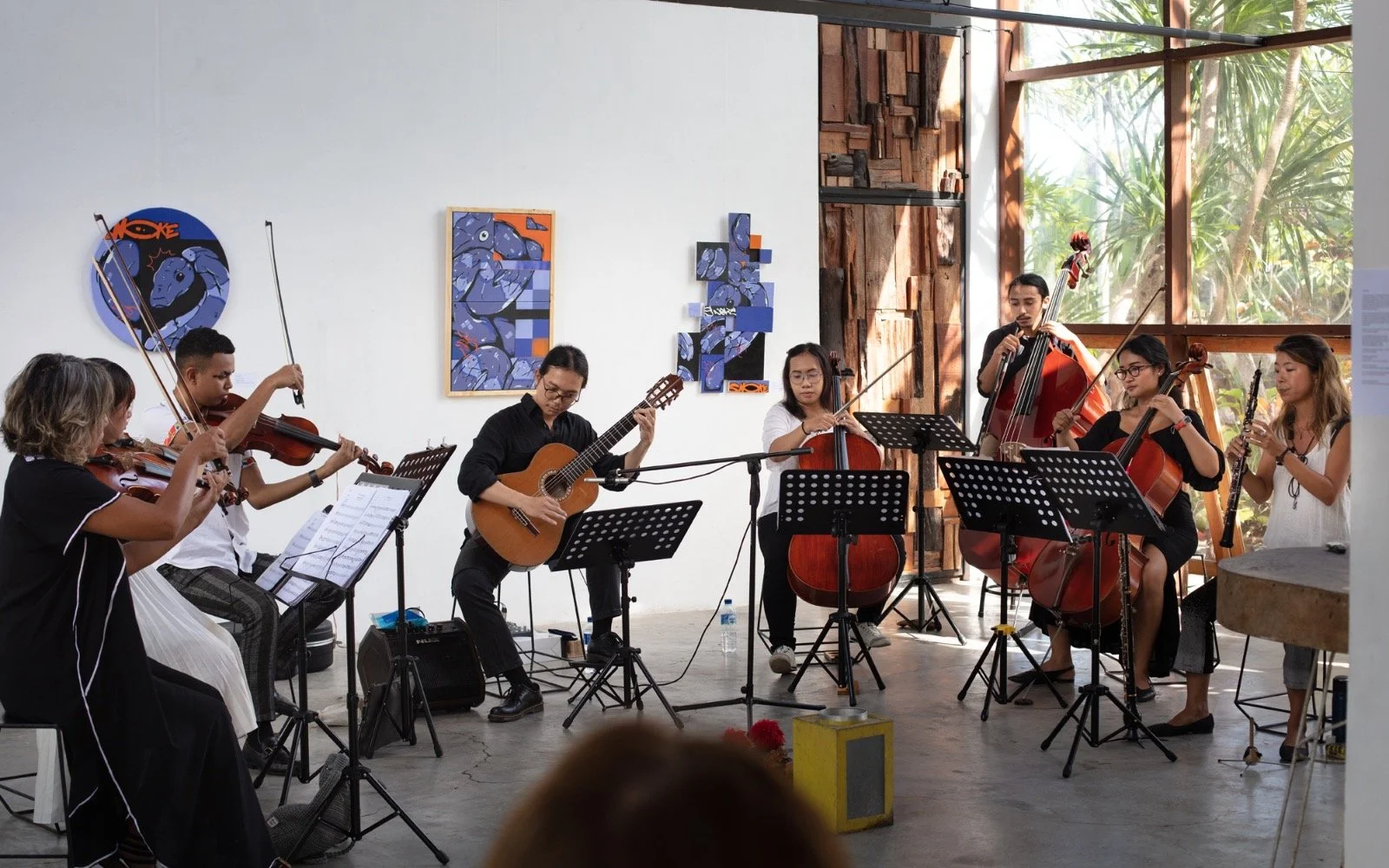 A group of musicians playing classical instruments during a performance in an art gallery with modern artwork on the wall and large windows showing trees outside.