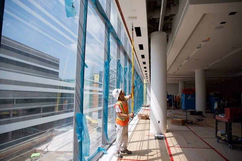 Construction worker wearing a helmet and safety vest cleaning large glass windows inside a building under construction or renovation.