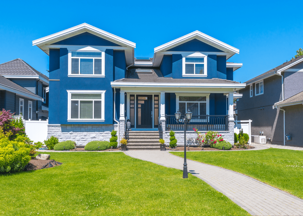 Blue two-story house with white trim, front porch, landscaped yard, and walking path under clear blue sky.