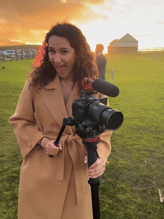 Woman with curly hair wearing a tan coat holding a professional video camera outdoors during sunset.