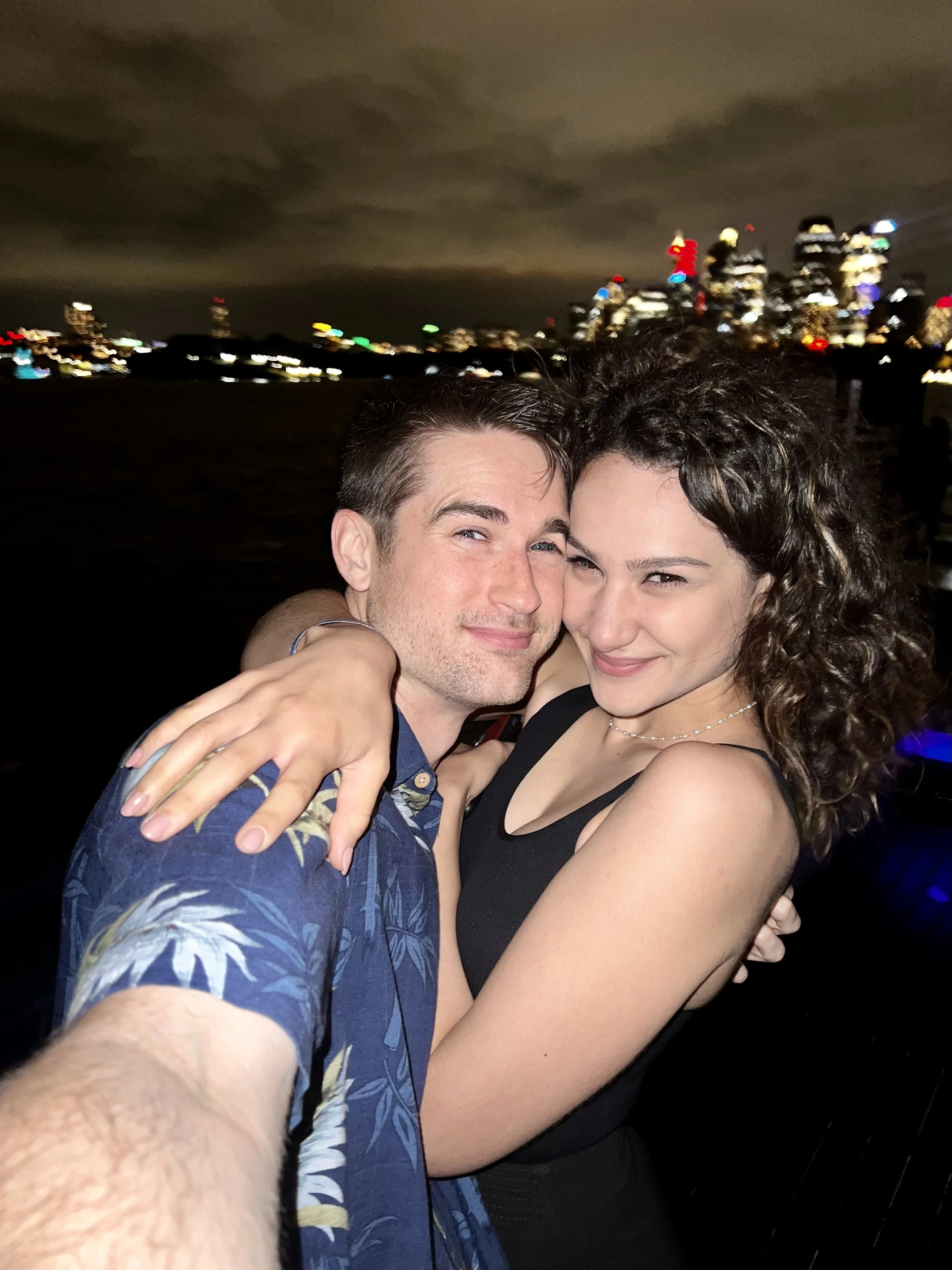 A young man and woman embrace for a selfie at night with a city skyline and water in the background.
