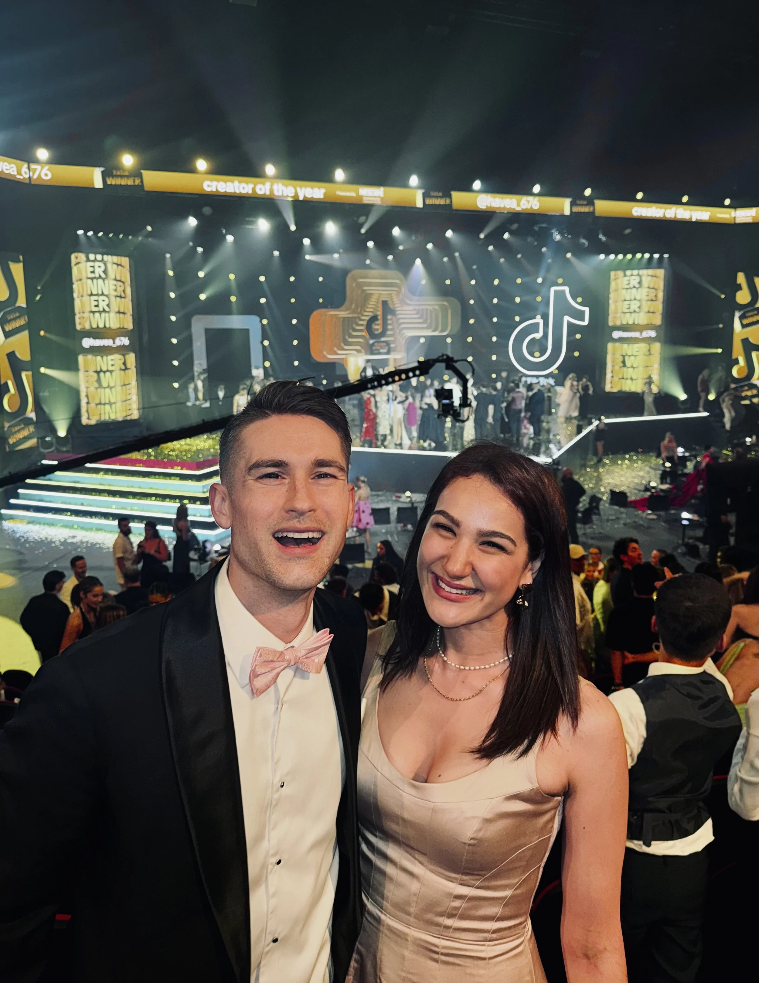 A man in a tuxedo with a pink bow tie and a woman in a beige dress posing together at an awards event, with a stage featuring TikTok and other social media logos in the background.