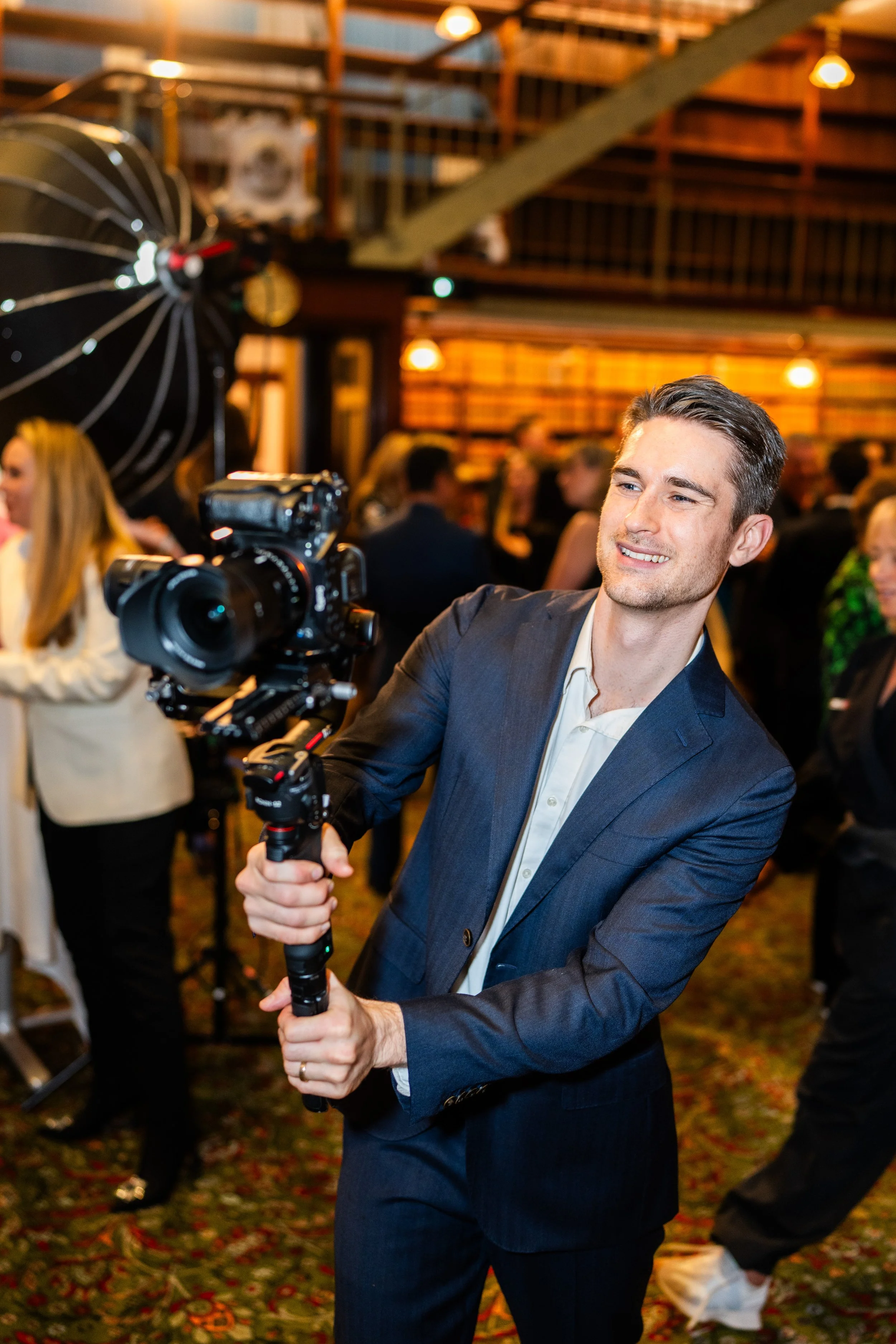 A man in a dark suit holding a camera on a stabilizer at a social gathering indoors with warm lighting.