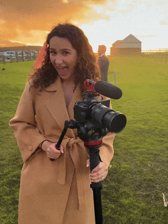 A woman with curly hair holding a professional camera with a microphone, smiling at sunset in an outdoor park.