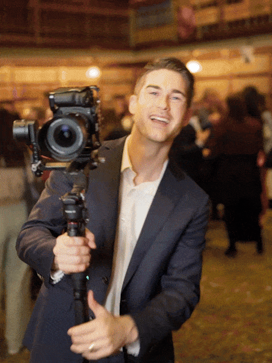 A smiling man in a dark suit holding a professional camera on a tripod at an indoor event.