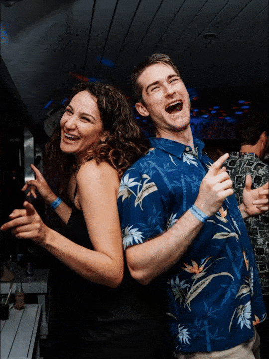 Two young adults, a woman with curly hair and a man with short hair, laughing and enjoying themselves at a party, standing close together in a dark indoor setting.