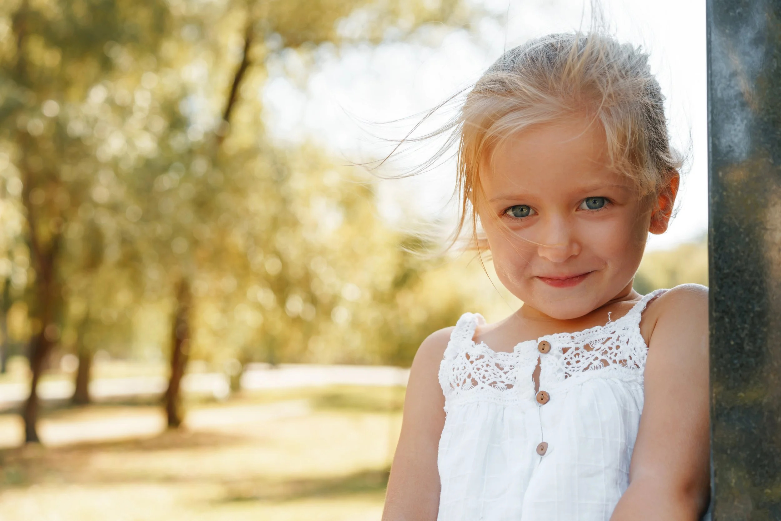 Ein junges Mädchen steht im Freien, trägt ein weißes Kleid mit Knöpfen und lächelt leicht, während es an einer dunklen Oberfläche lehnt. Im Hintergrund sind Bäume mit gelblichen Blättern bei sonnigem Wetter zu sehen.