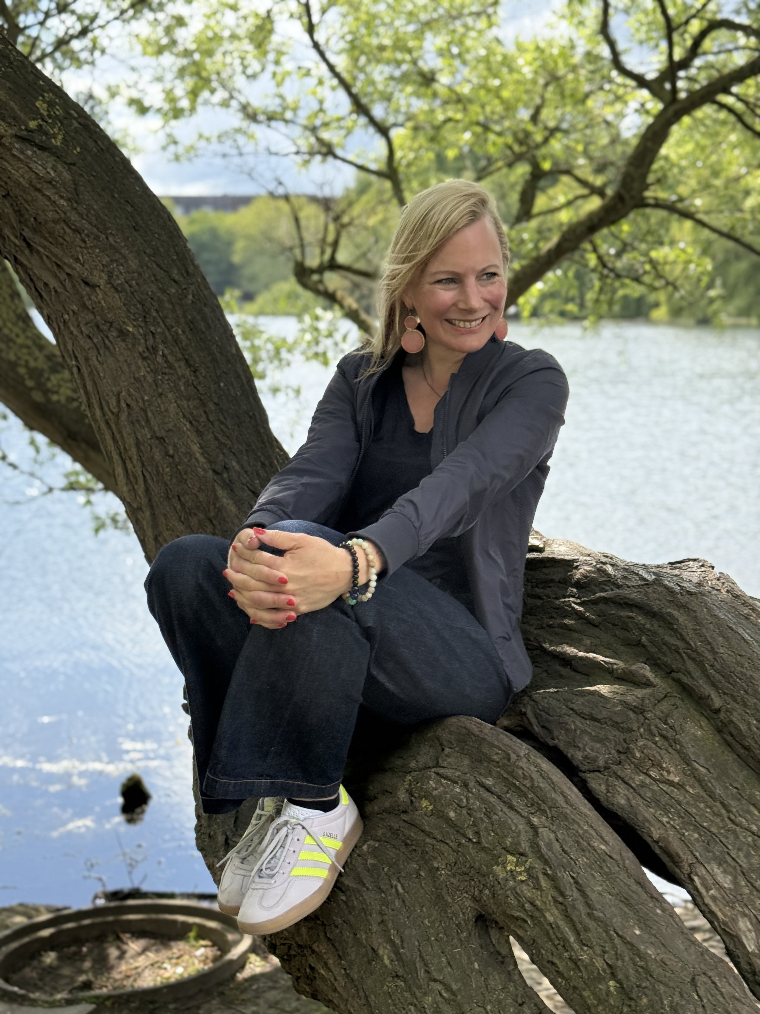 Birgit Elke sitzt auf einem Baumstamm im Grünen, neben einem Fluss, trägt eine dunkle Jacke, jeans und weiße Turnschuhe, trägt Ohrringe und Armbänder, umgeben von Blättern und Wasser.