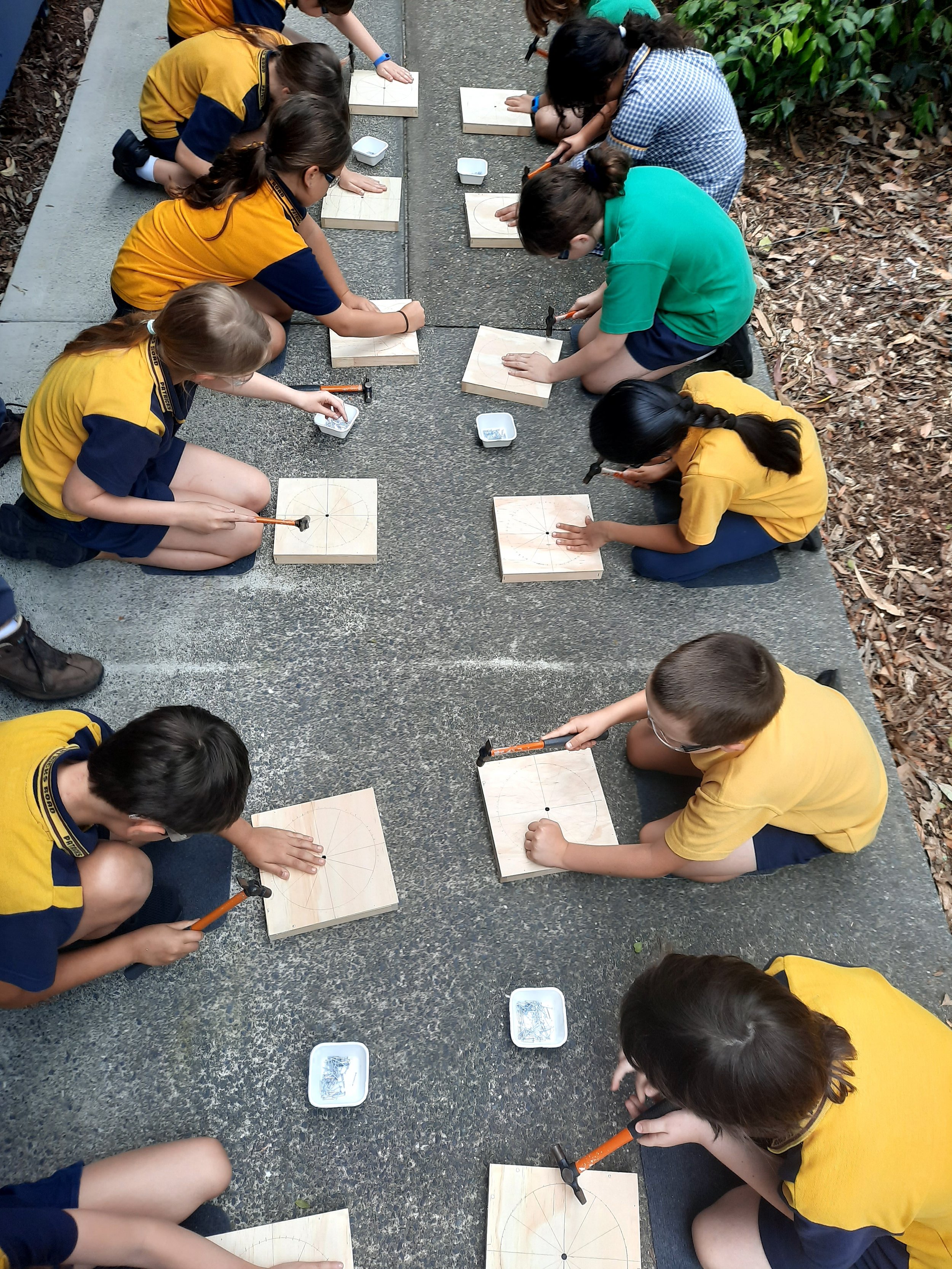 Children in school uniforms are sitting on the ground, using hammers and chisels to carve and shape wooden squares, possibly engaged in a woodworking activity outdoors.