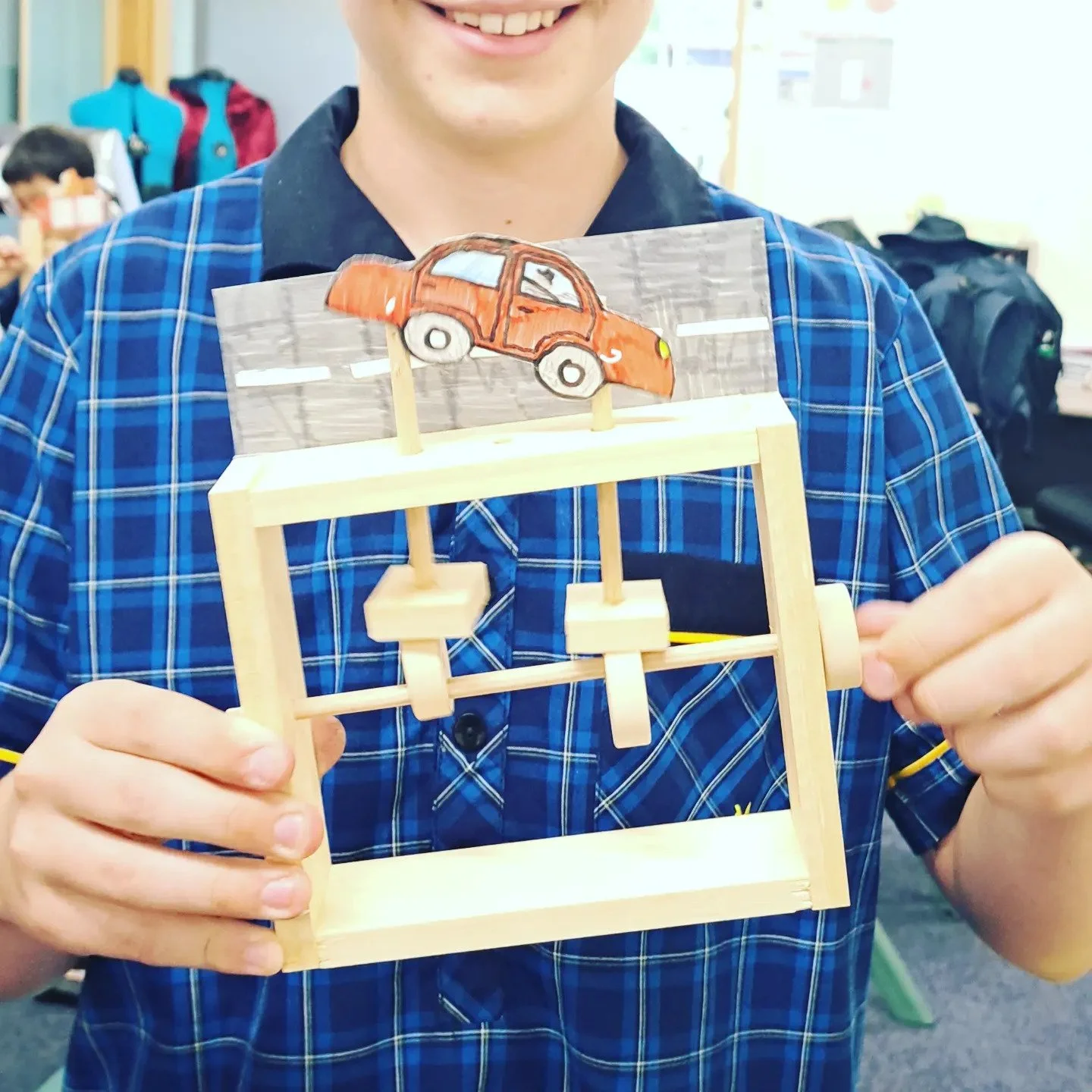 Person holding a wooden swing with a hand-drawn orange car on a stick atop it, in a classroom setting.