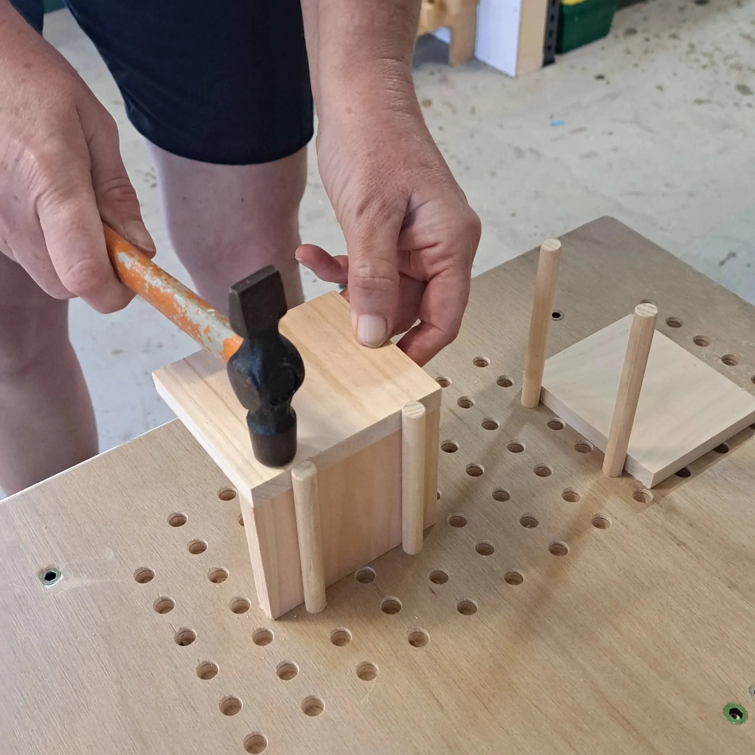 Person hammering a small wooden block on a workbench with wood dowels and holes.