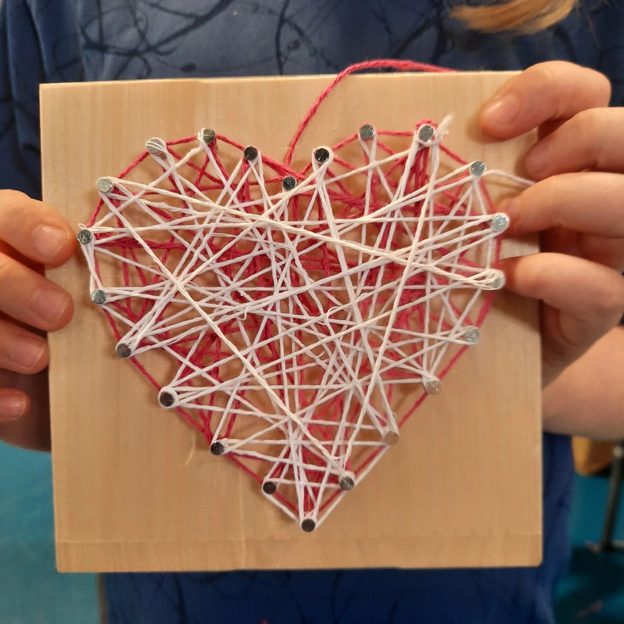 Hands holding a wooden square board with a red and white string art heart design secured with nails.