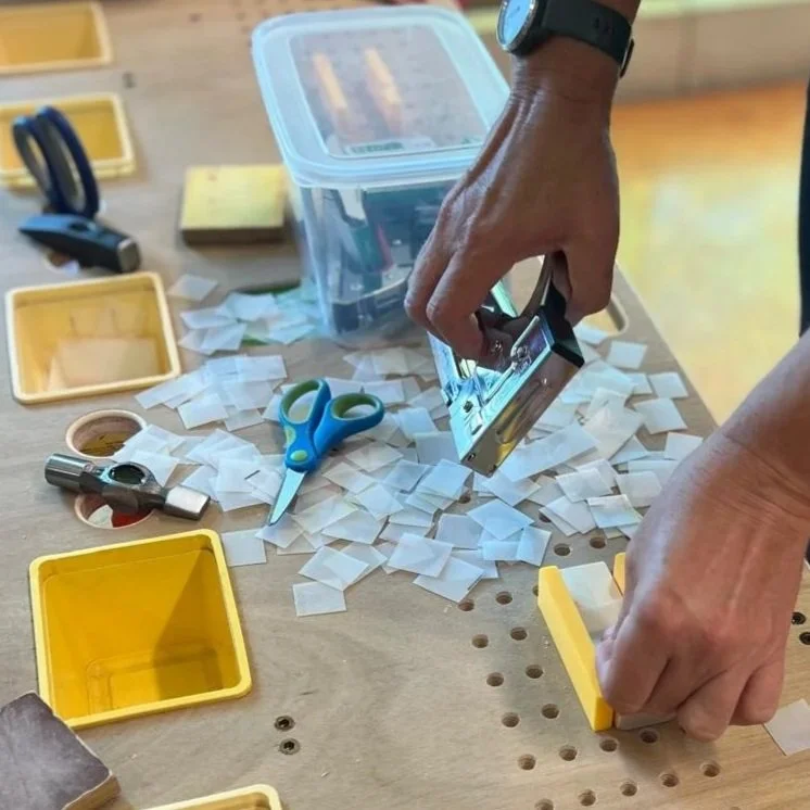 Person using a paper punch to cut small paper pieces on a cluttered work surface with scissors, tape, and storage containers.