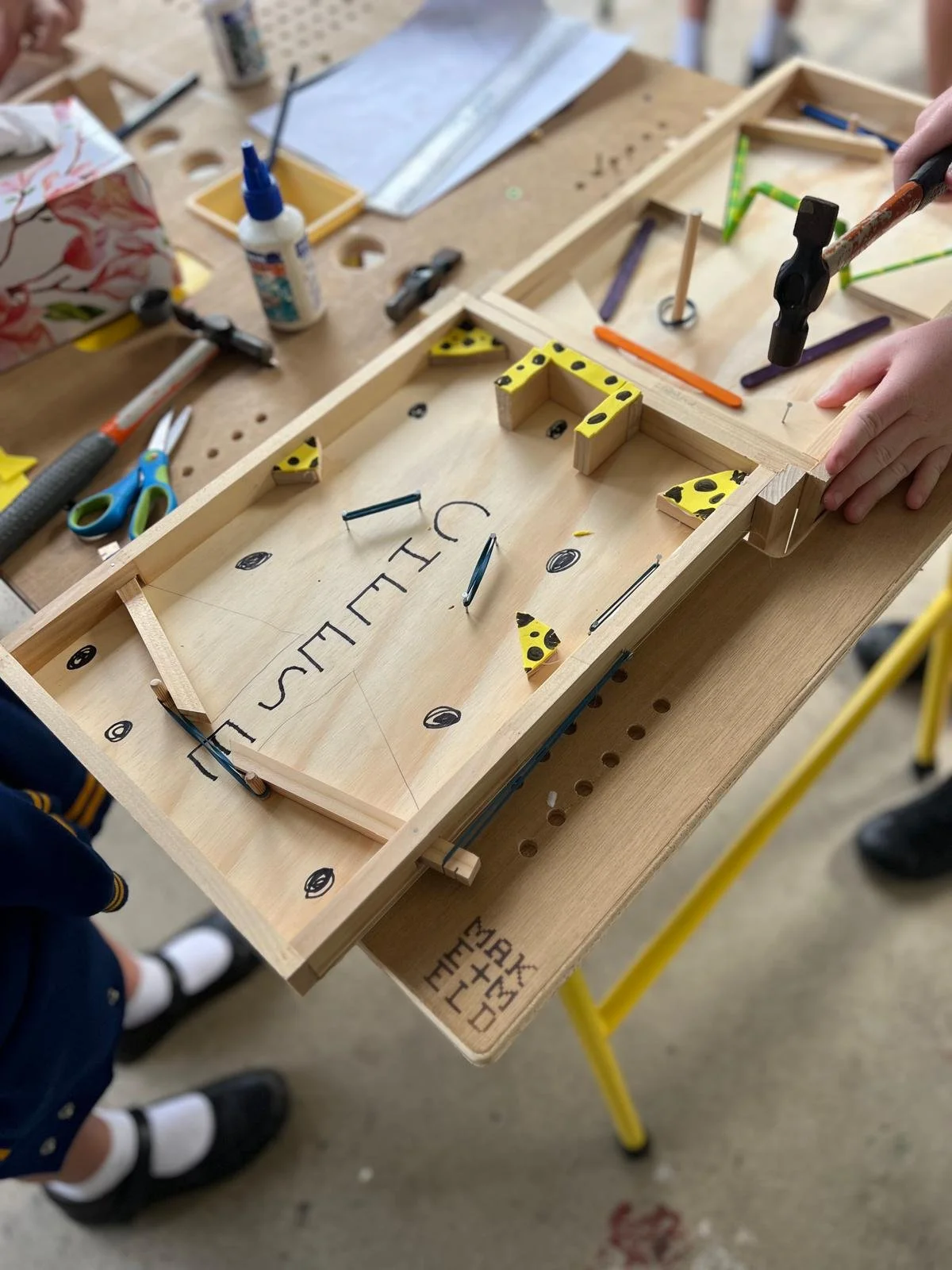 A wooden pinball machine with yellow bumpers, black dots, and colorful ramps, being assembled by a person in a workshop.