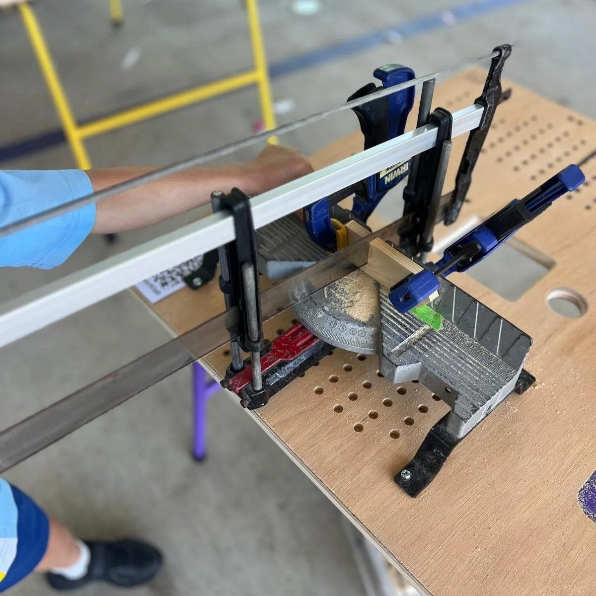 A woodworking setup with clamps securing a piece of wood on a workbench, a square, and a saw, in a workshop environment.