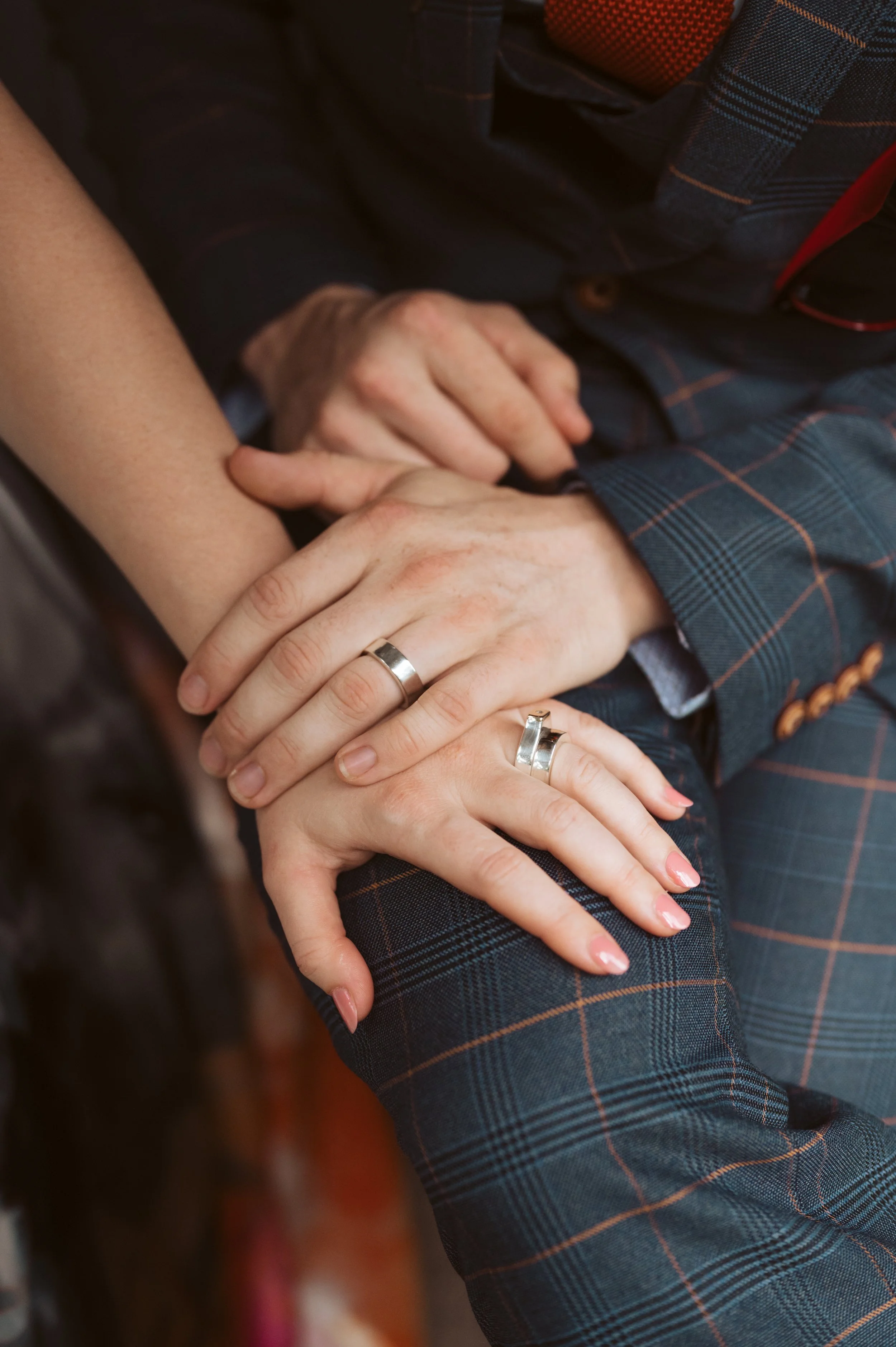 Two people wearing matching silver rings on their ring fingers, holding hands. One person is wearing a plaid suit, and the other is wearing a short-sleeve top with a tan skin tone.