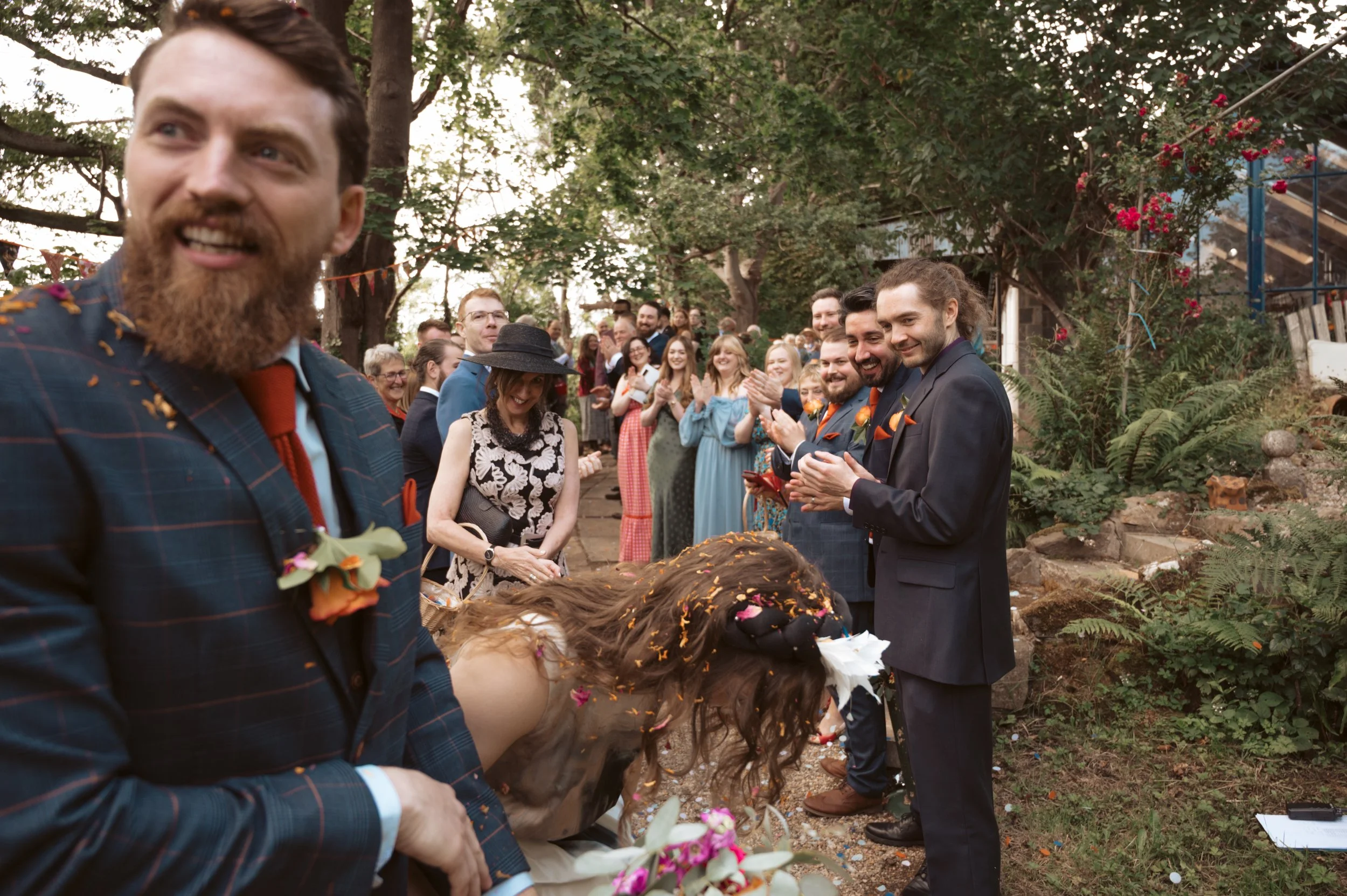 A group of wedding guests outdoors, clapping and smiling, with a bride bowing with her head down, in a garden setting with trees and flowers.