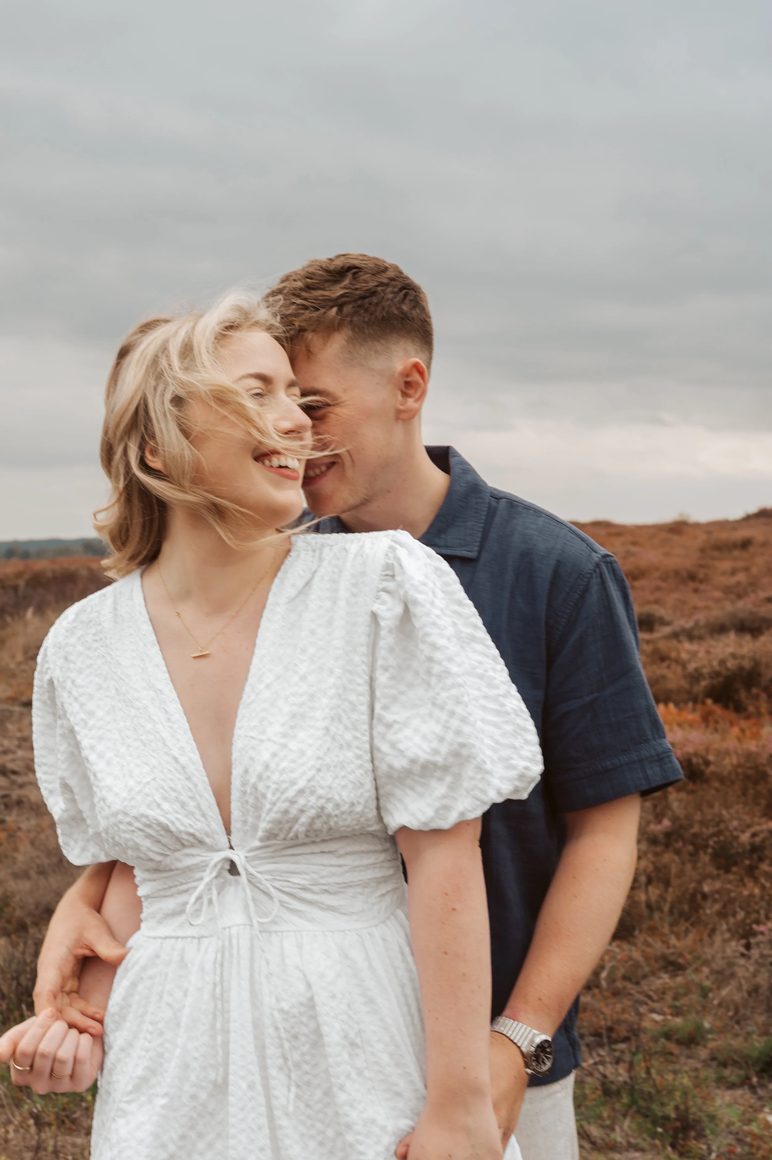 A happy couple laughing and embracing outdoors on a moody day during their engagement shoot,  with a landscape of brownish shrubbery in the background.