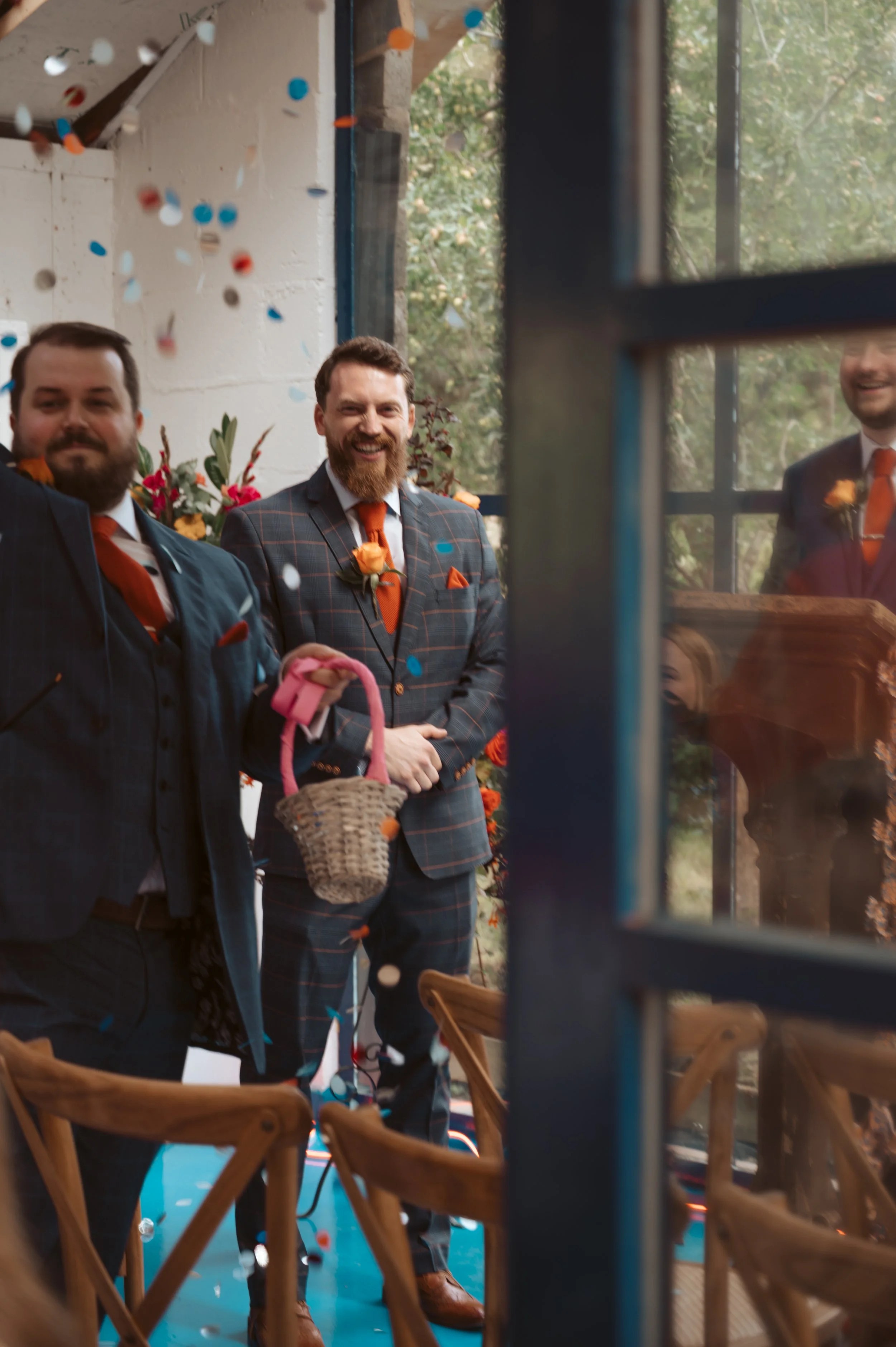 Celebrants at a wedding reception celebrating with confetti, three men in suits with orange ties and boutonnières, one holding a pink basket, inside an industrial space with large windows and wooden chairs.