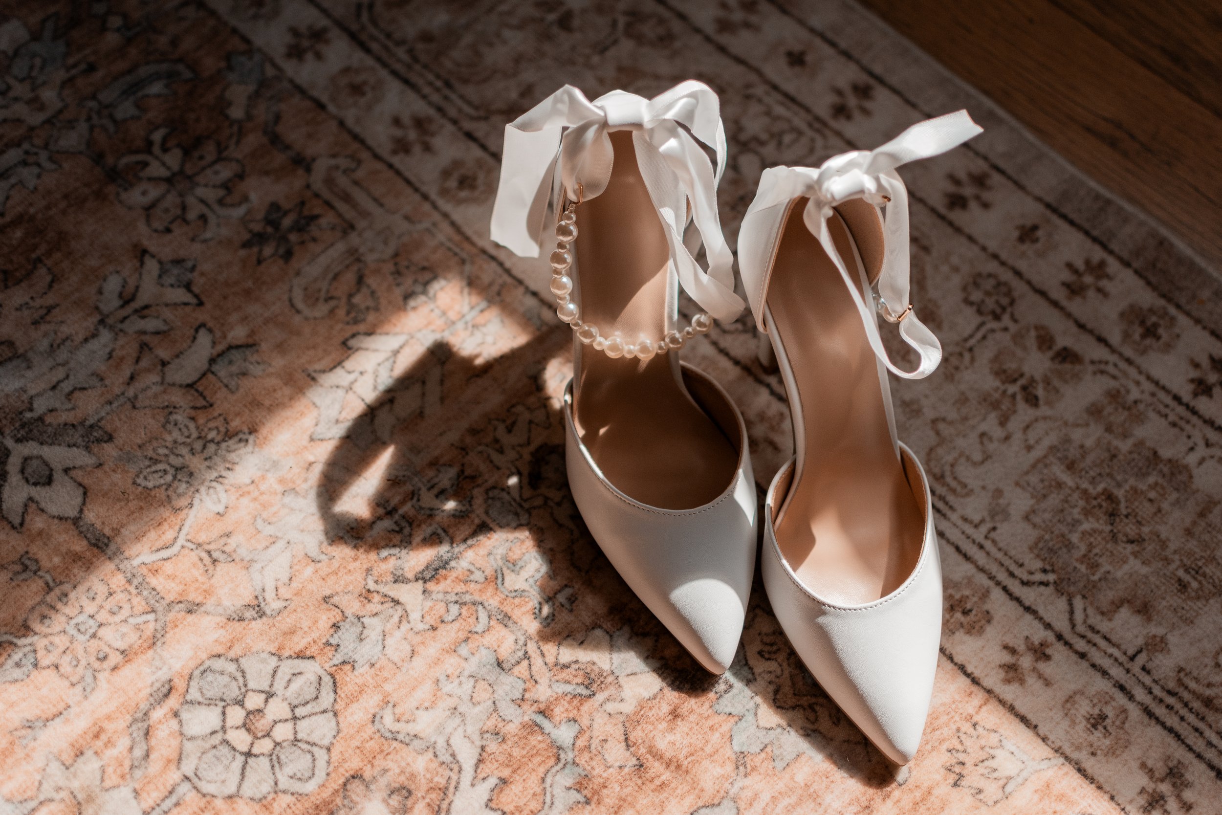 A pair of white high-heeled shoes with ribbon ties on an ornate rug, with one shoe adorned with a pearl anklet.