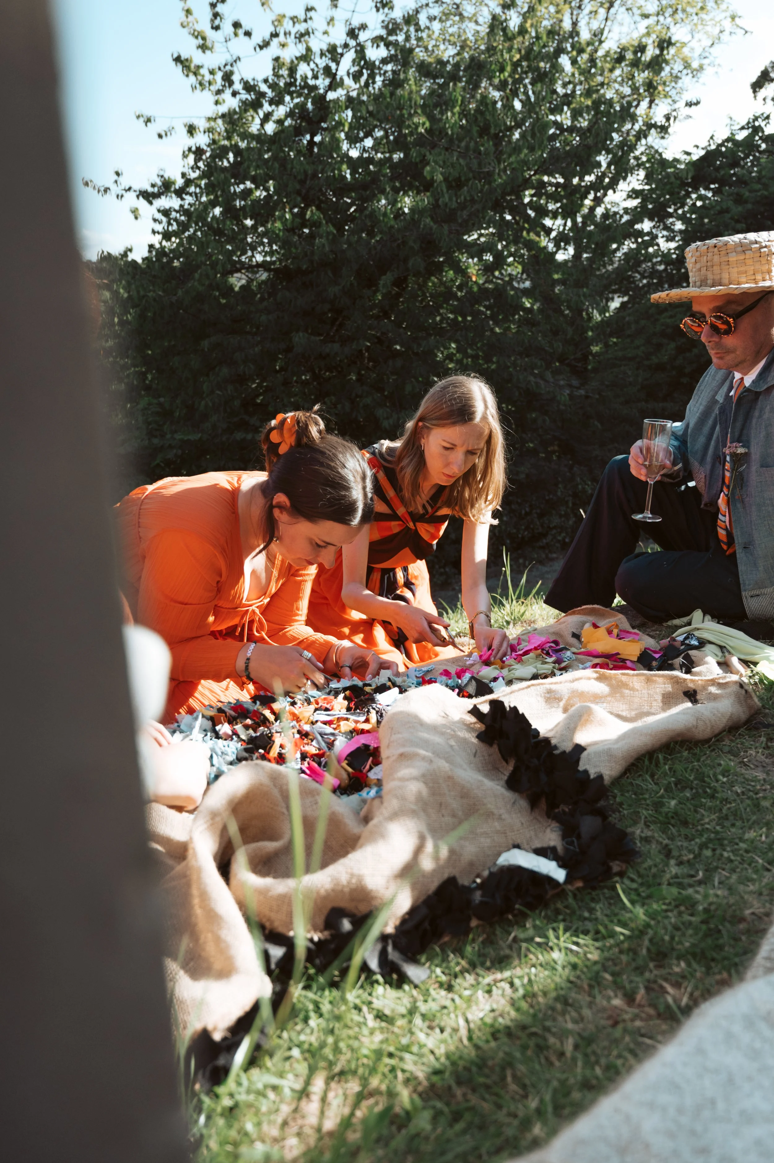 People sitting on the grass sifting through colorful fabric scraps during an outdoor gathering on a sunny day.