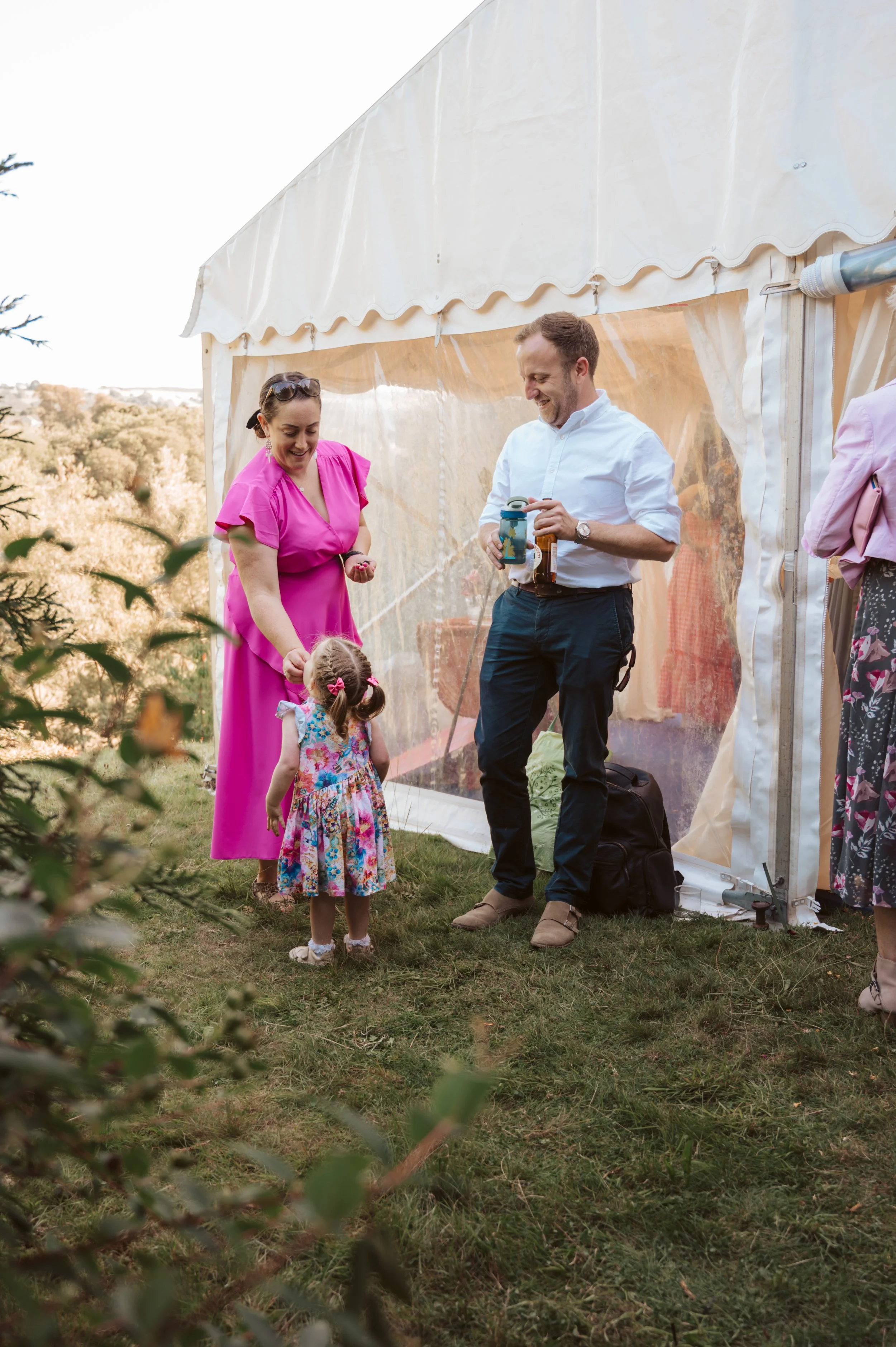 A woman in a pink dress, a man, and a young girl with braided hair and a floral dress enjoying a moment outside a white tent, with greenery and trees in the background.