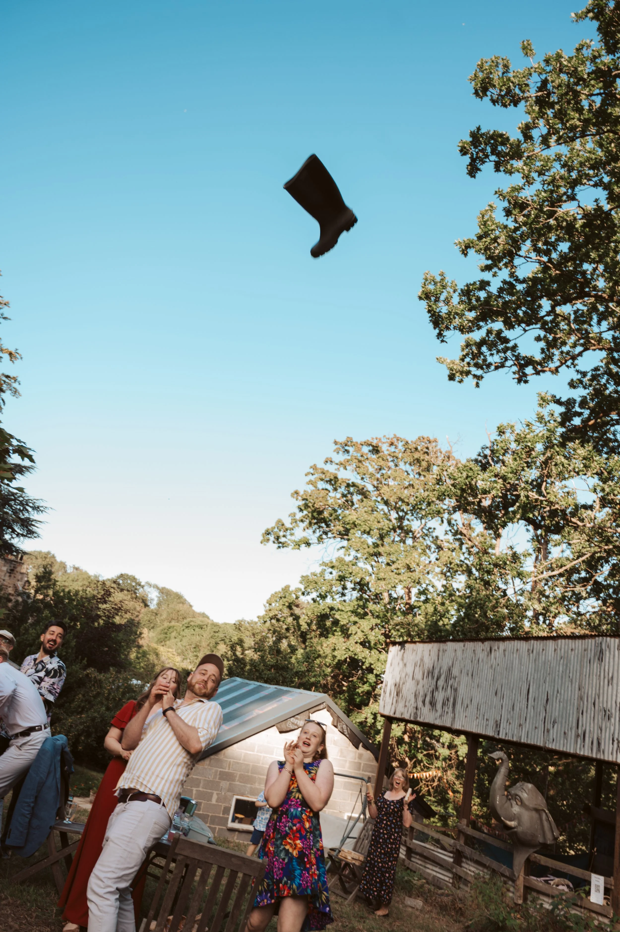 People watching a black boot fly through the air outdoors, with trees and a small building in the background.