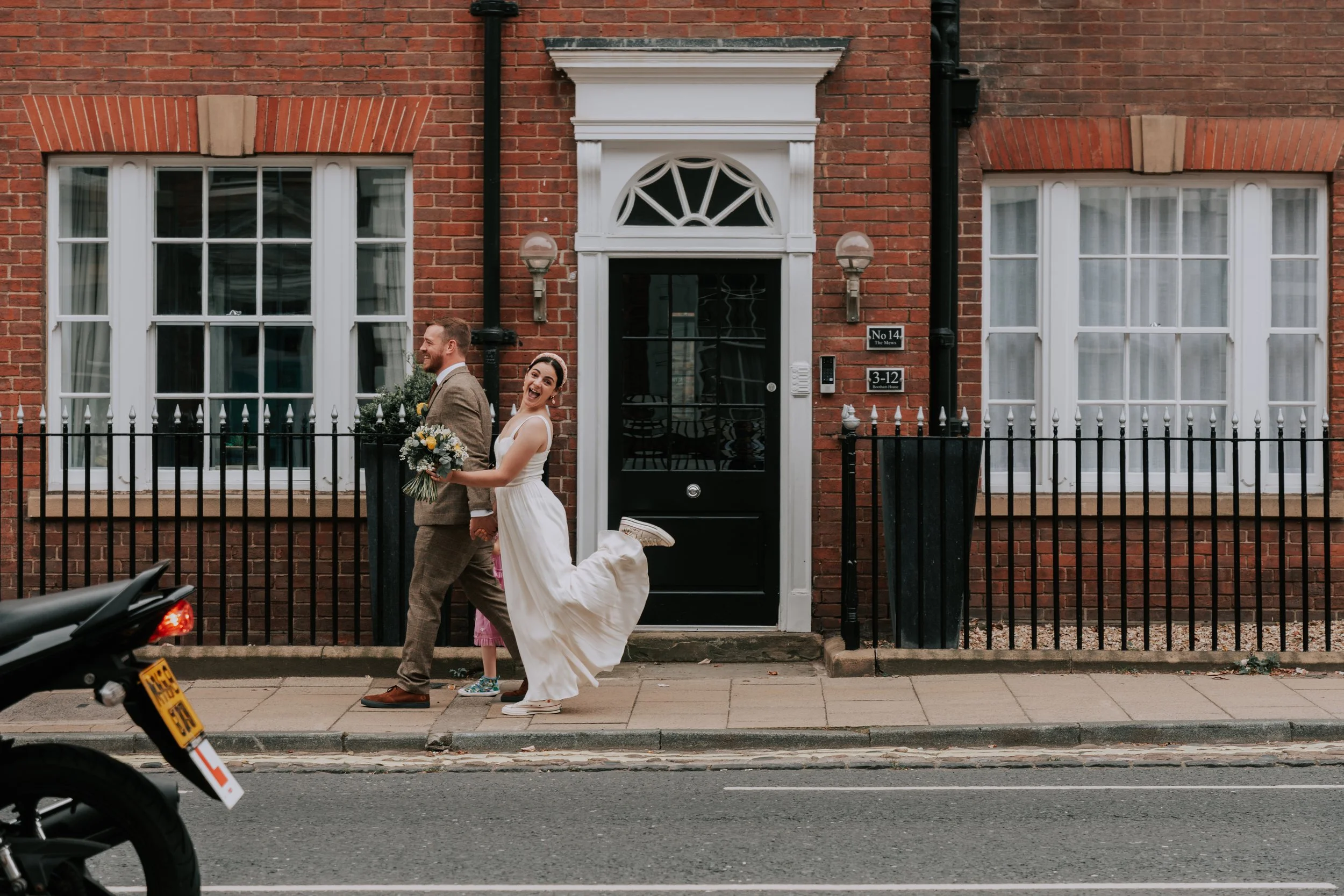 A newlywed couple walking on a sidewalk outside a brick house, with the bride playfully holding up her dress and the groom looking ahead and smiling. The bride is holding a bouquet of flowers.
