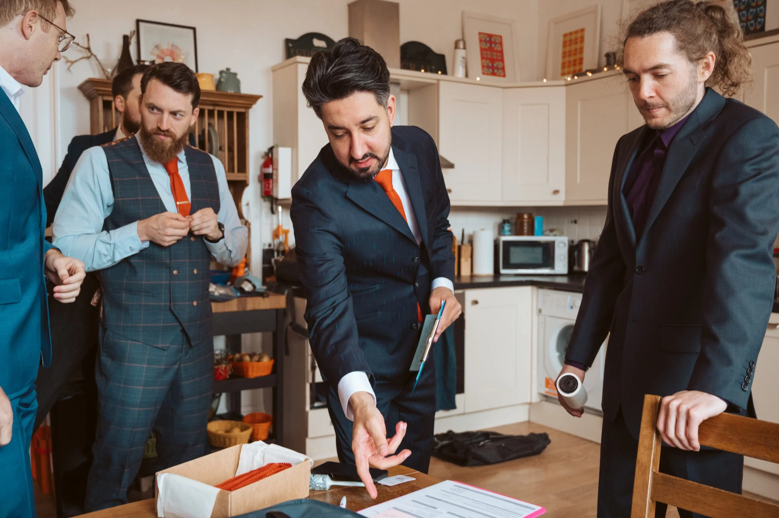 A group of men in suits gathered around a table in a kitchen, with one man showing something on a clipboard to the others.