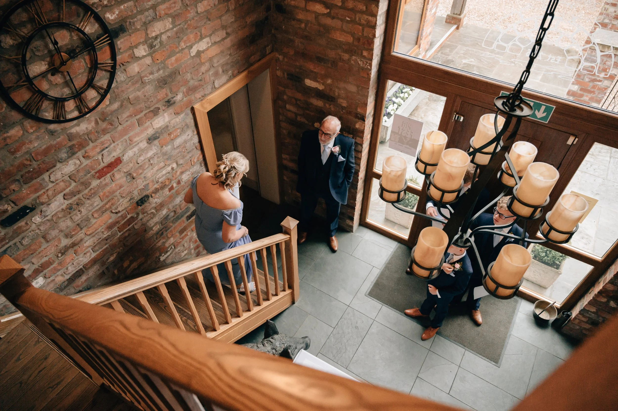 View from above of a wedding reception or formal gathering inside a brick-walled building. Four people are near the entrance; one woman in a light blue dress is standing on a staircase, while three men in suits are standing near the door. A large cha