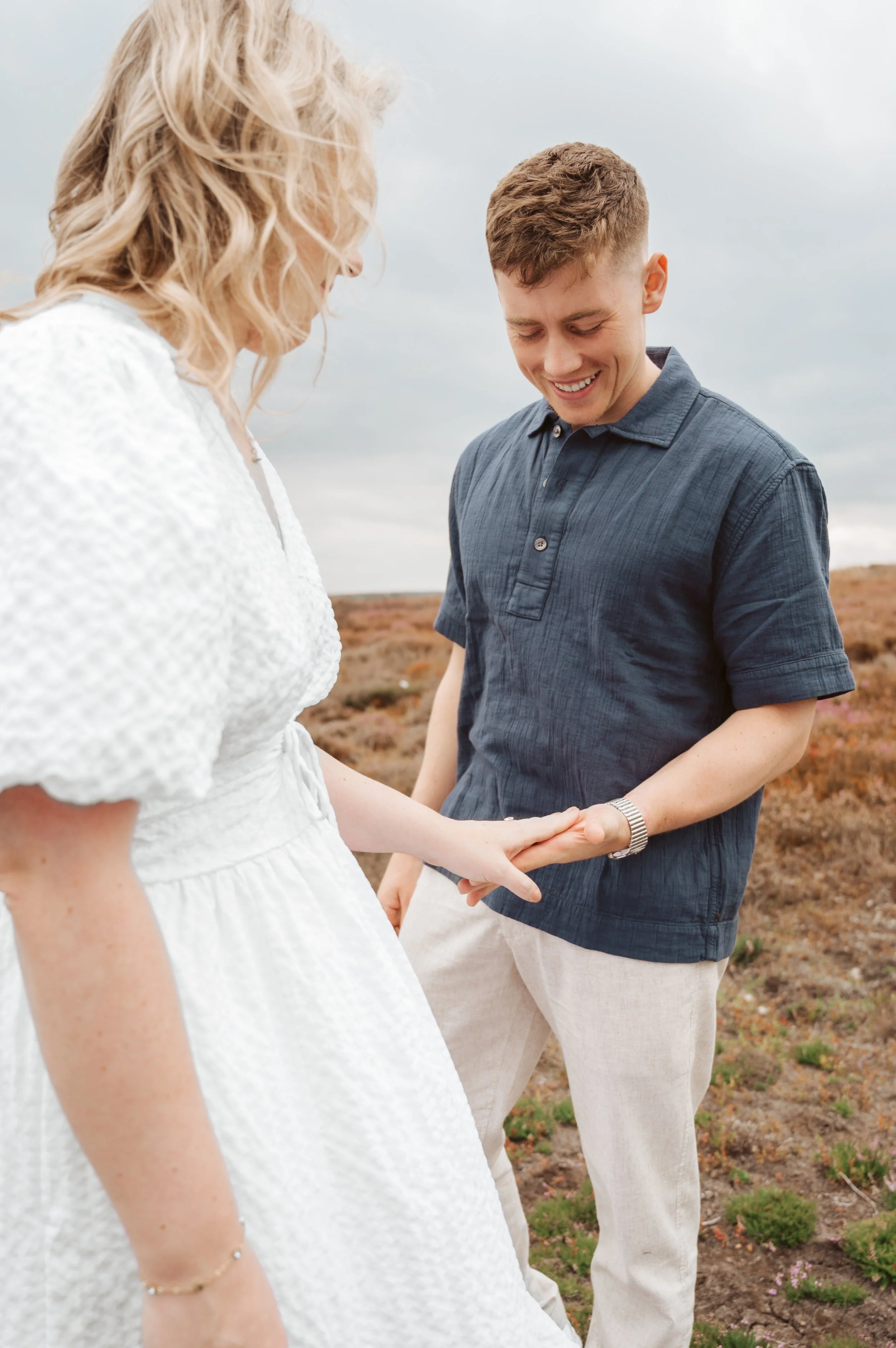 A young man and woman standing outdoors in a natural landscape, holding hands and smiling at each other.