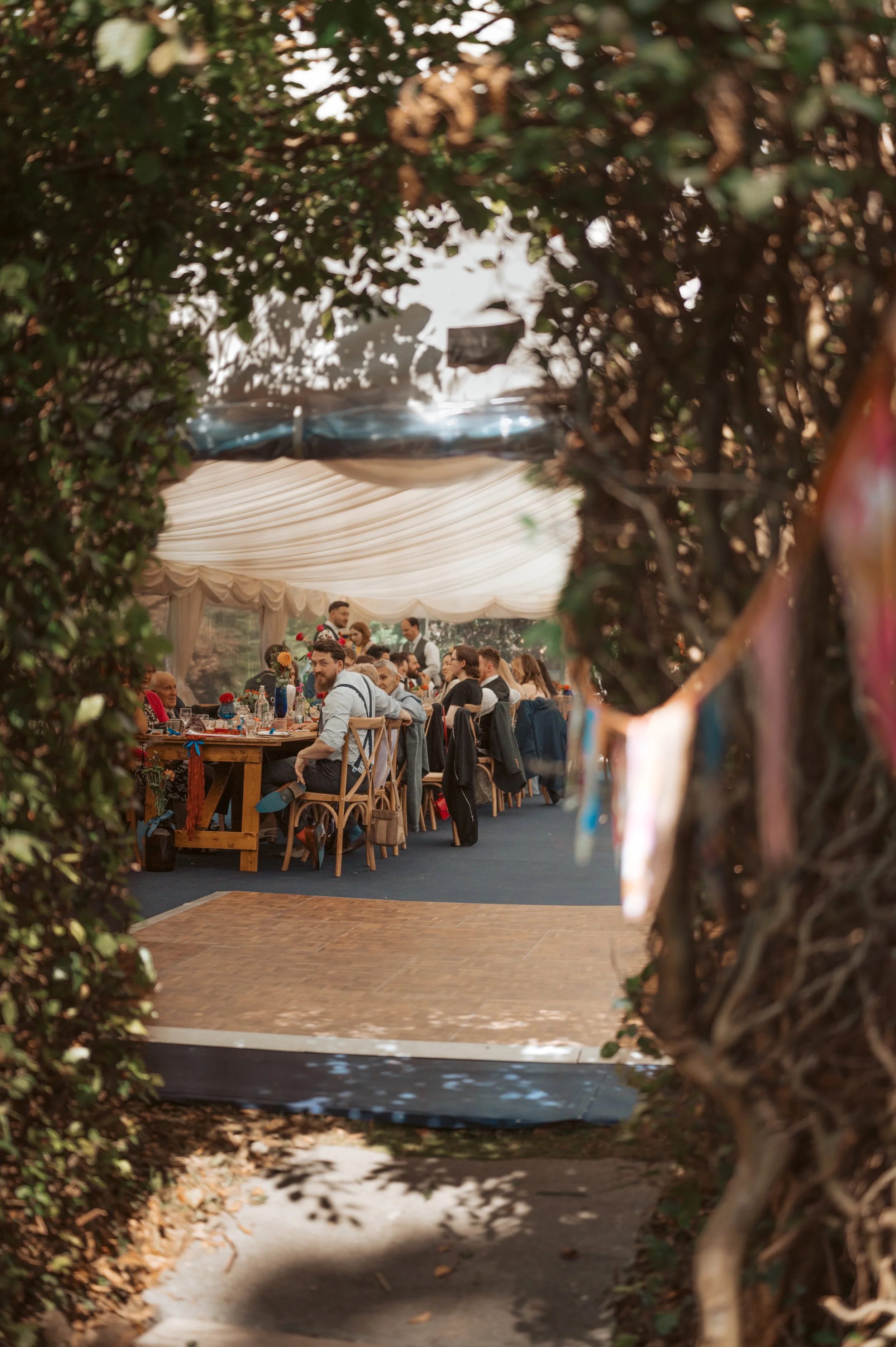 People gathered around a banquet table at an outdoor event, seen through a natural frame of foliage and trees.