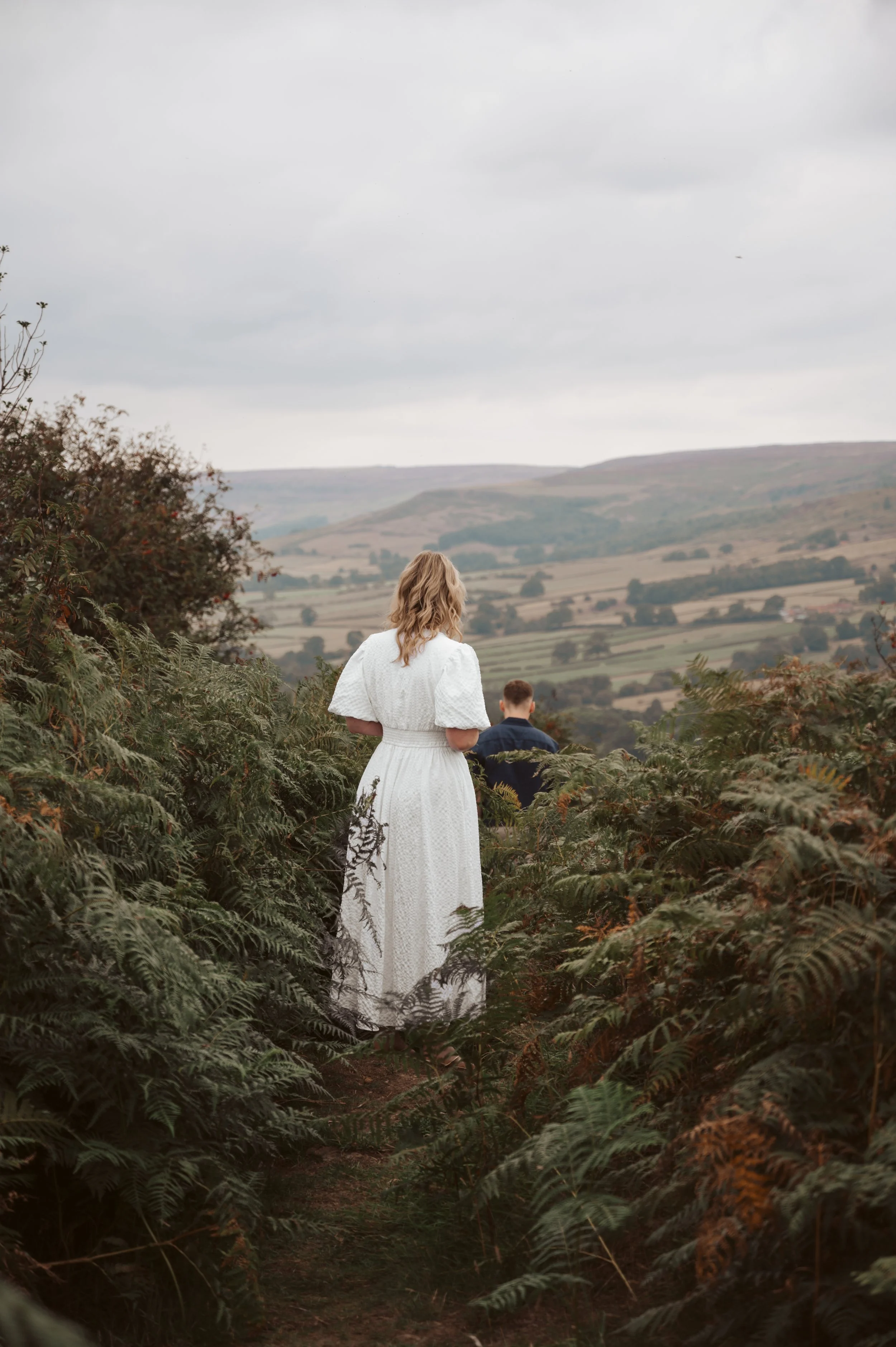 A woman in a white dress standing on a bushy trail, looking at a man in the distance, with a rolling hilly landscape under a cloudy sky.