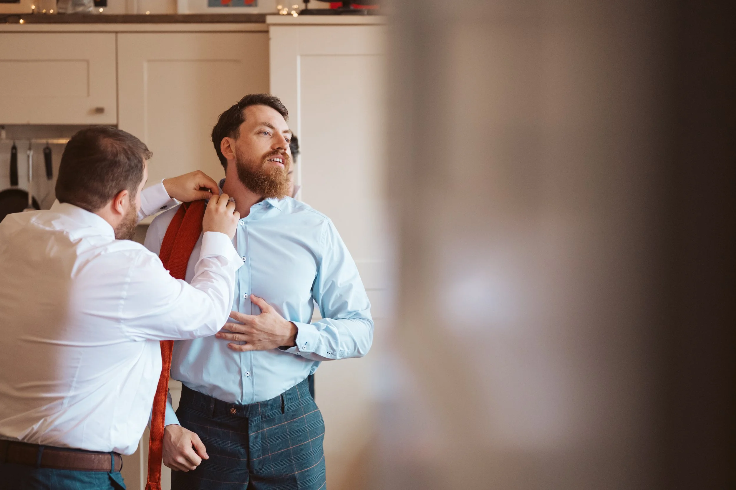 Two men in a kitchen, one helping the other adjust a red bag over his shoulder, with a focus on the man in a light blue shirt.