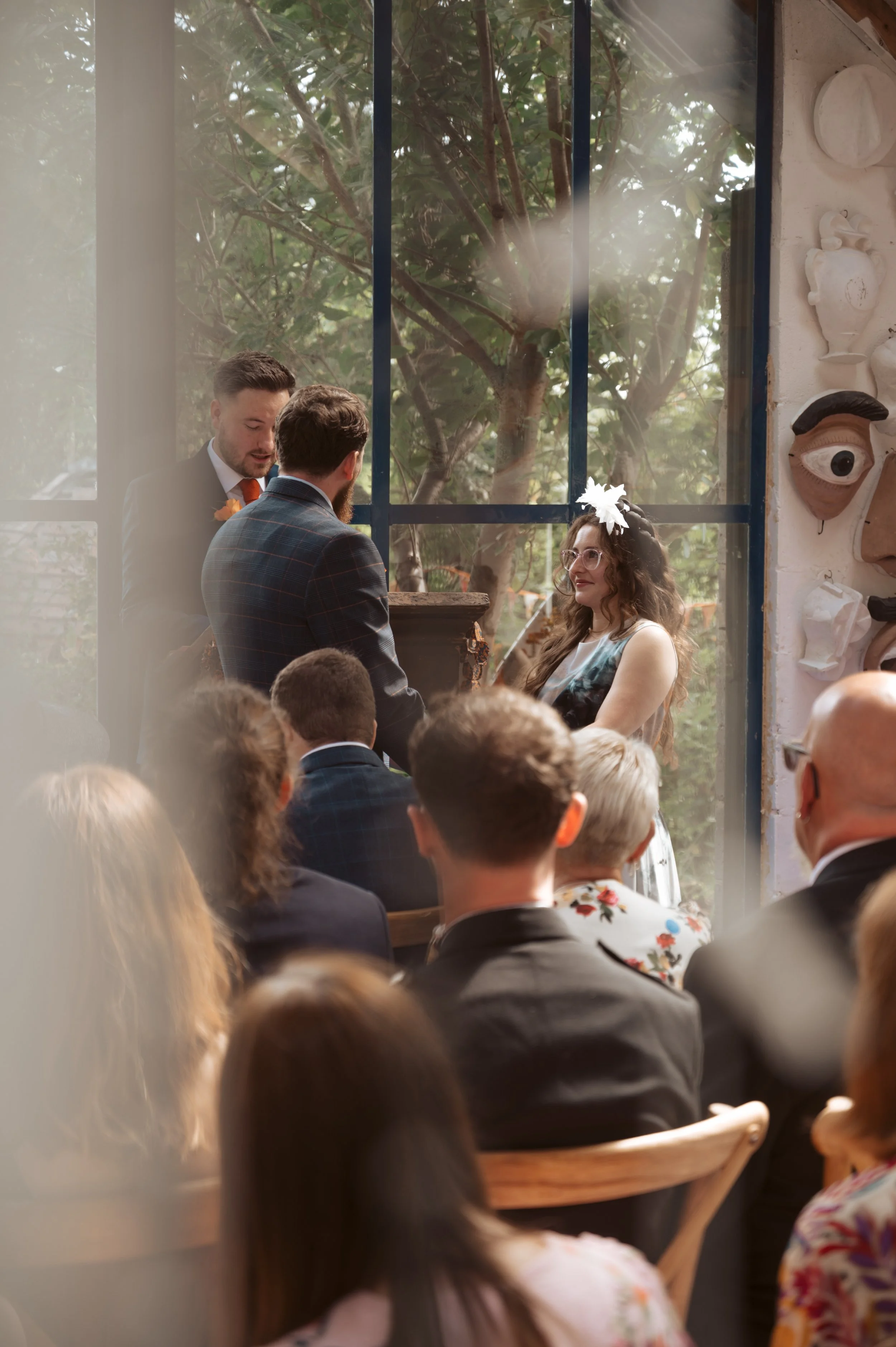 A wedding ceremony taking place indoors with a group of people seated, an officiant and groom standing, and a woman with glasses and a flower headpiece exchanging vows in front of a large window with trees outside.