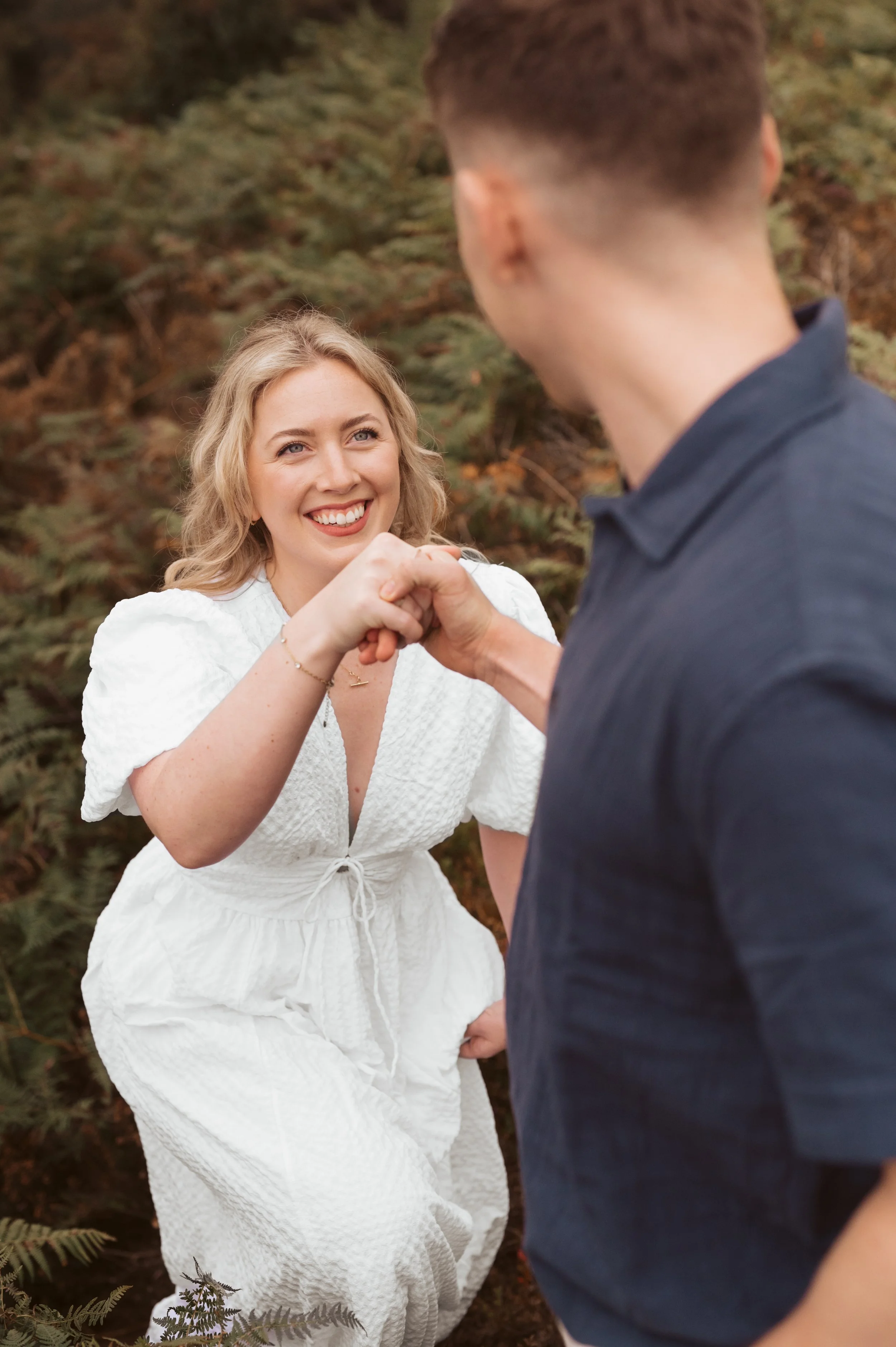 A woman in a white dress smiling and holding hands with a man in a dark shirt, outdoors with green foliage in the background.
