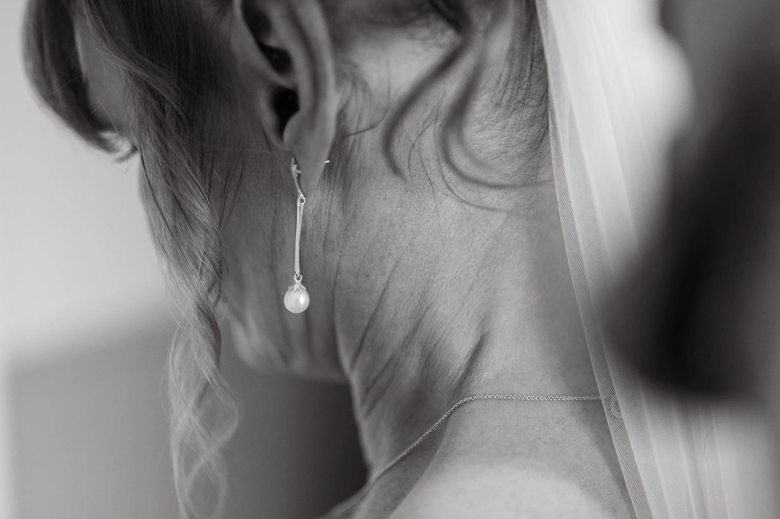 Close-up of a woman's neck and shoulder showing elegant pearl earrings and a delicate chain necklace, with a sheer fabric veil in the background, black and white photograph.