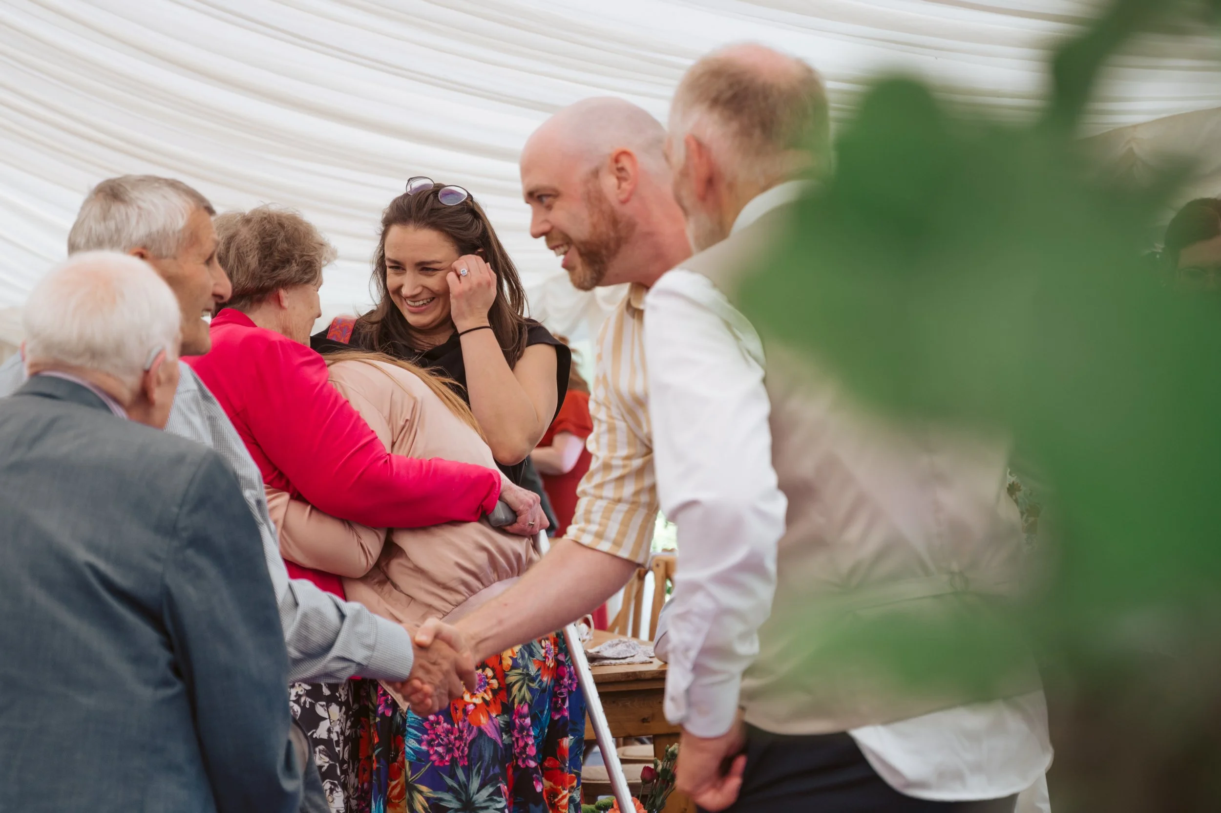People gathered at a celebration, hugging and shaking hands, smiling and laughing under a white tent.