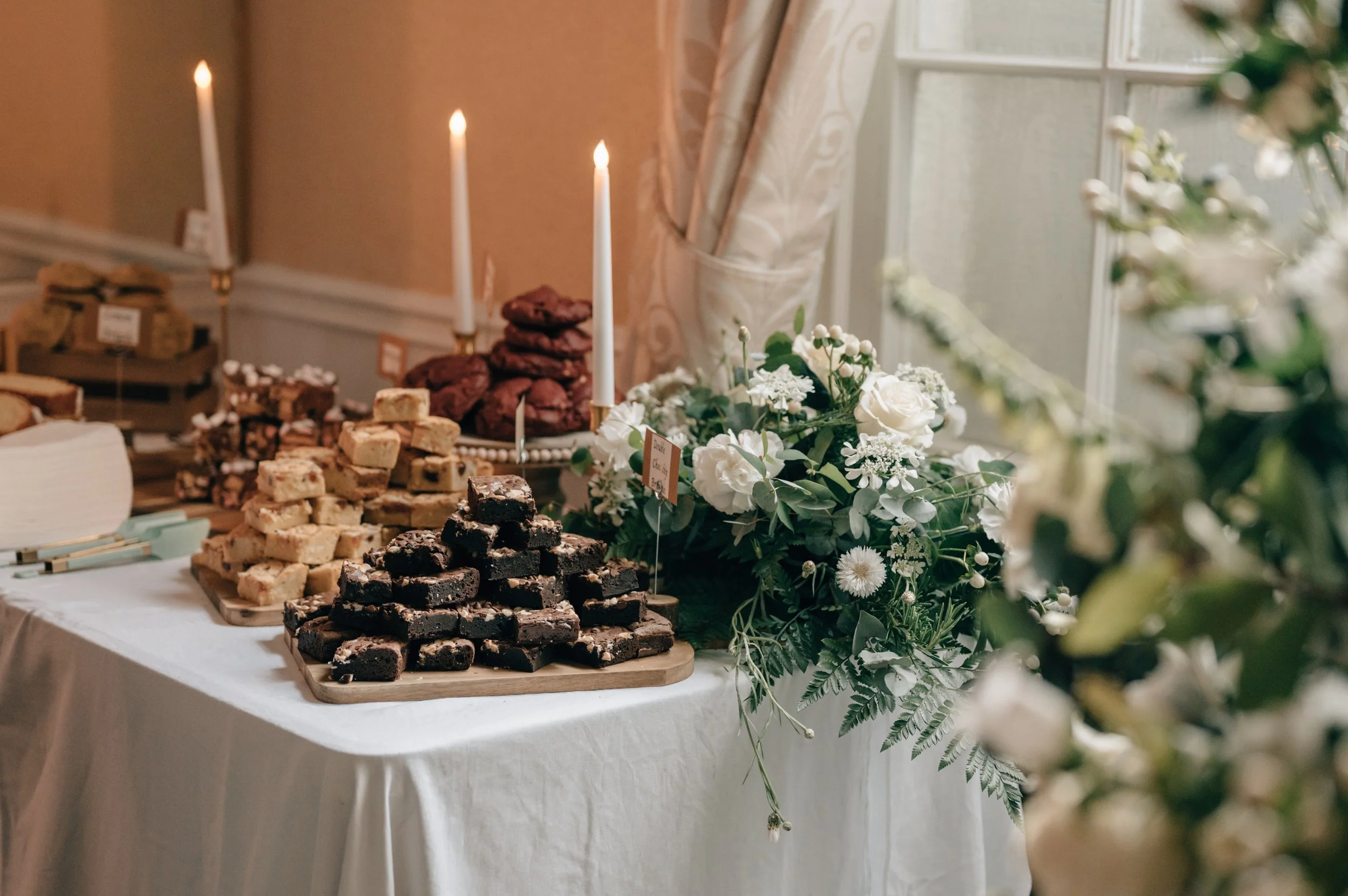 Dessert table with stacked brownies and blondies, white flowers, candles, and natural light from a window.