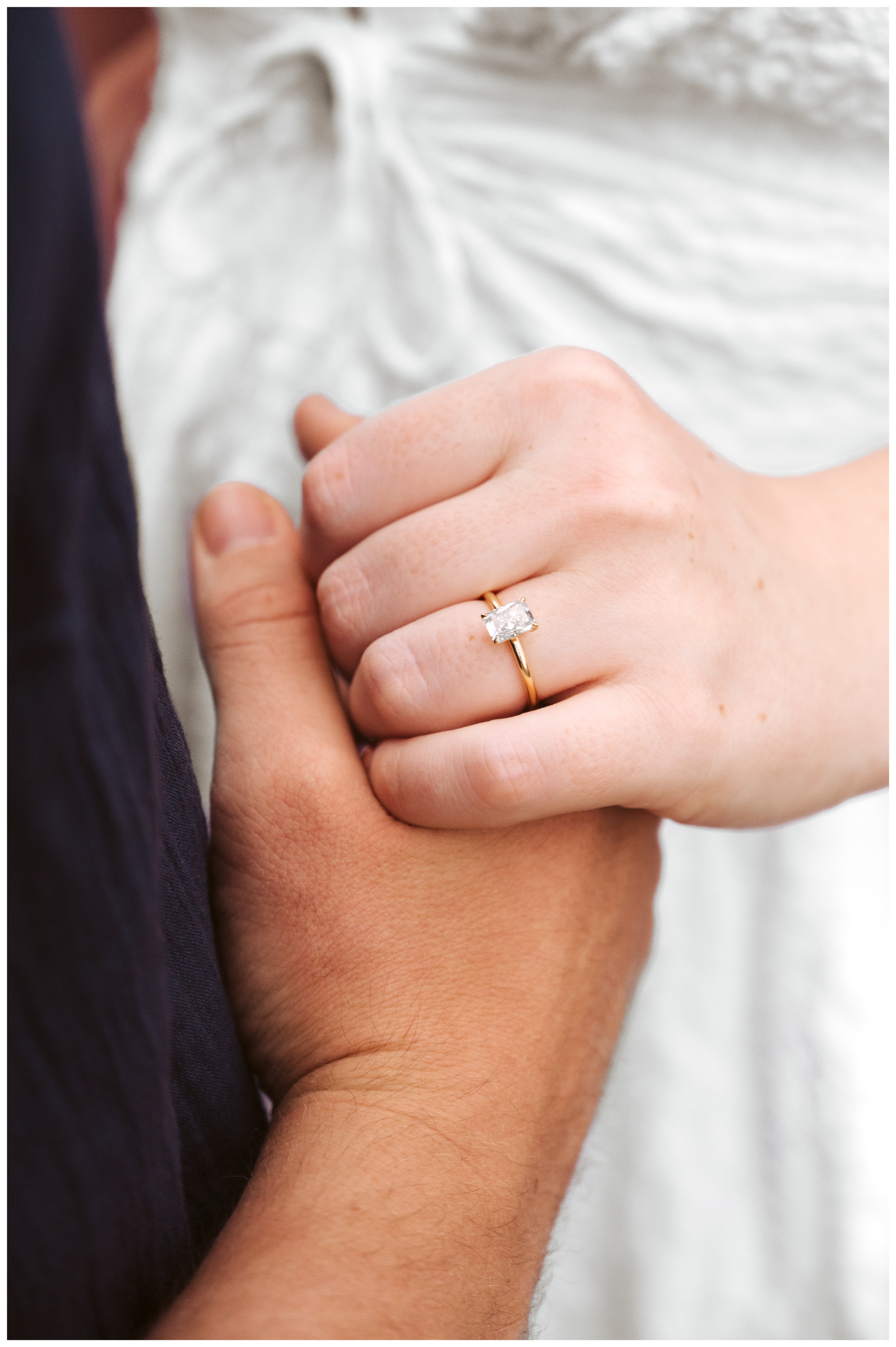 Close-up of a couple holding hands, the woman's hand with a gold ring with a large square diamond, the man's hand underneath.