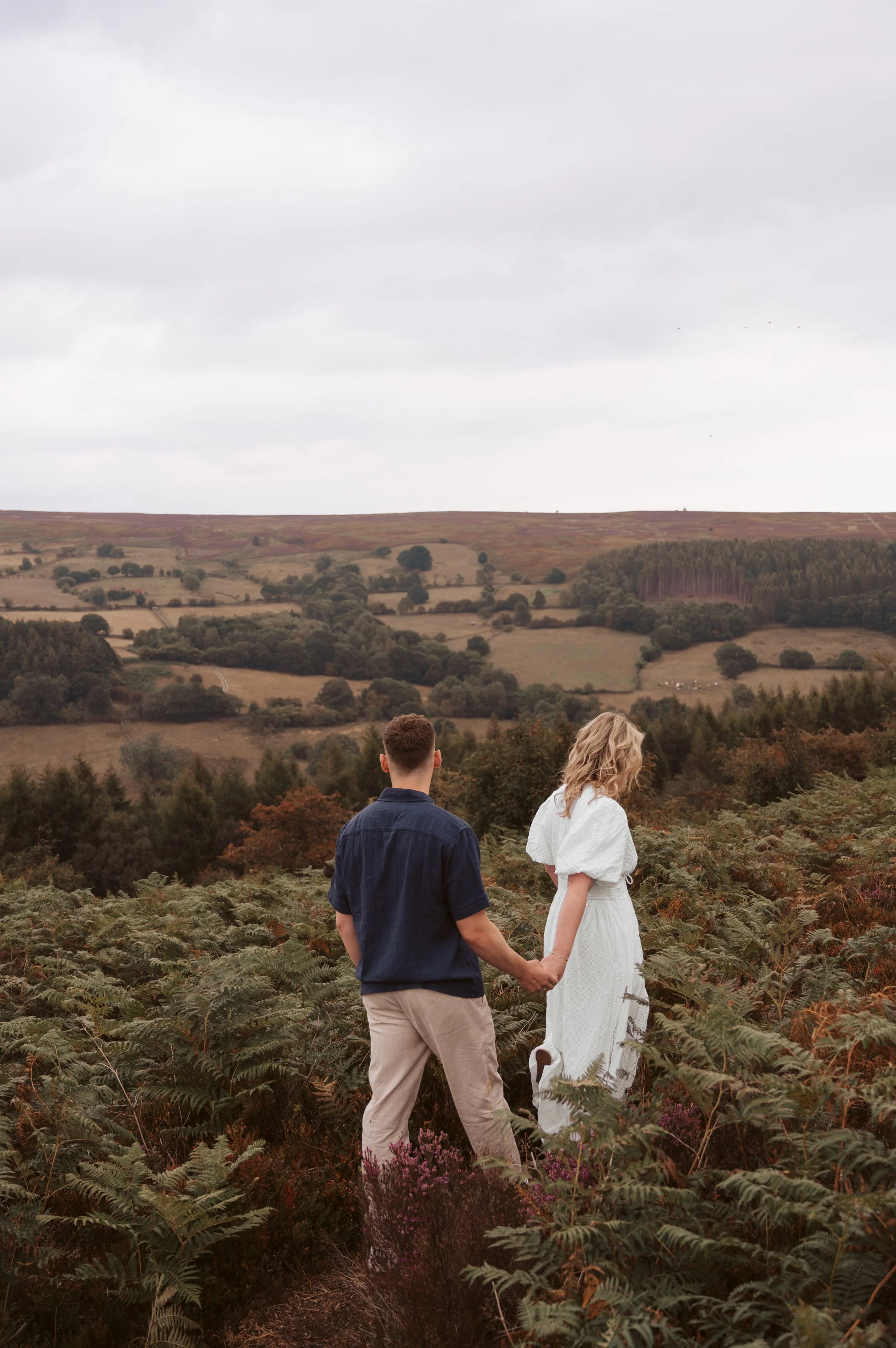 A couple holding hands, walking through a lush green field with a hilly countryside in the background under a cloudy sky.